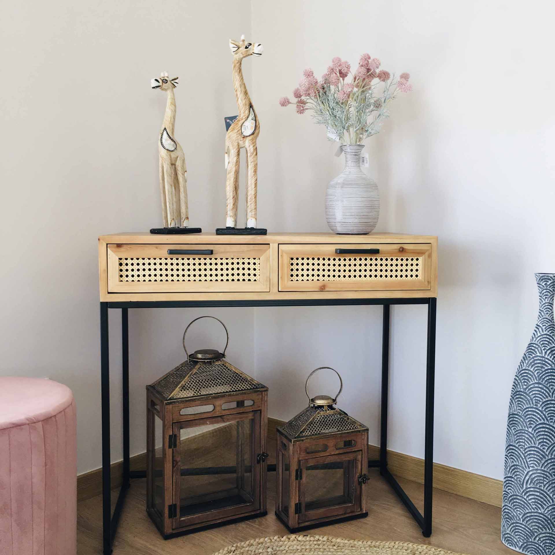 Consola em madeira clara e metal preto com gavetas e objetos decorativos, incluindo esculturas de girafas, vaso com flores e lanternas em chão de madeira