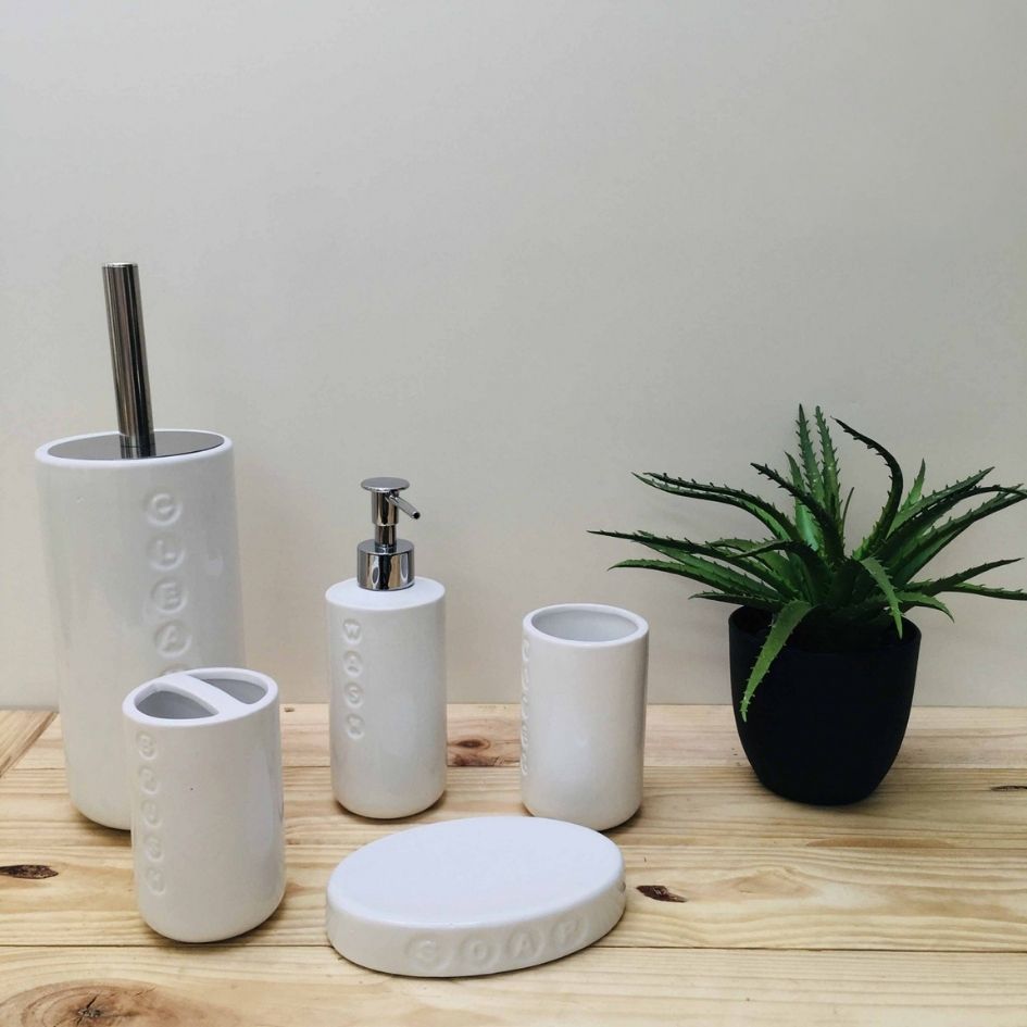 Conjunto de acessórios de casa-de-banho brancos em cerâmica com texto em relevo, sobre tábua de madeira com planta em vaso preto.
