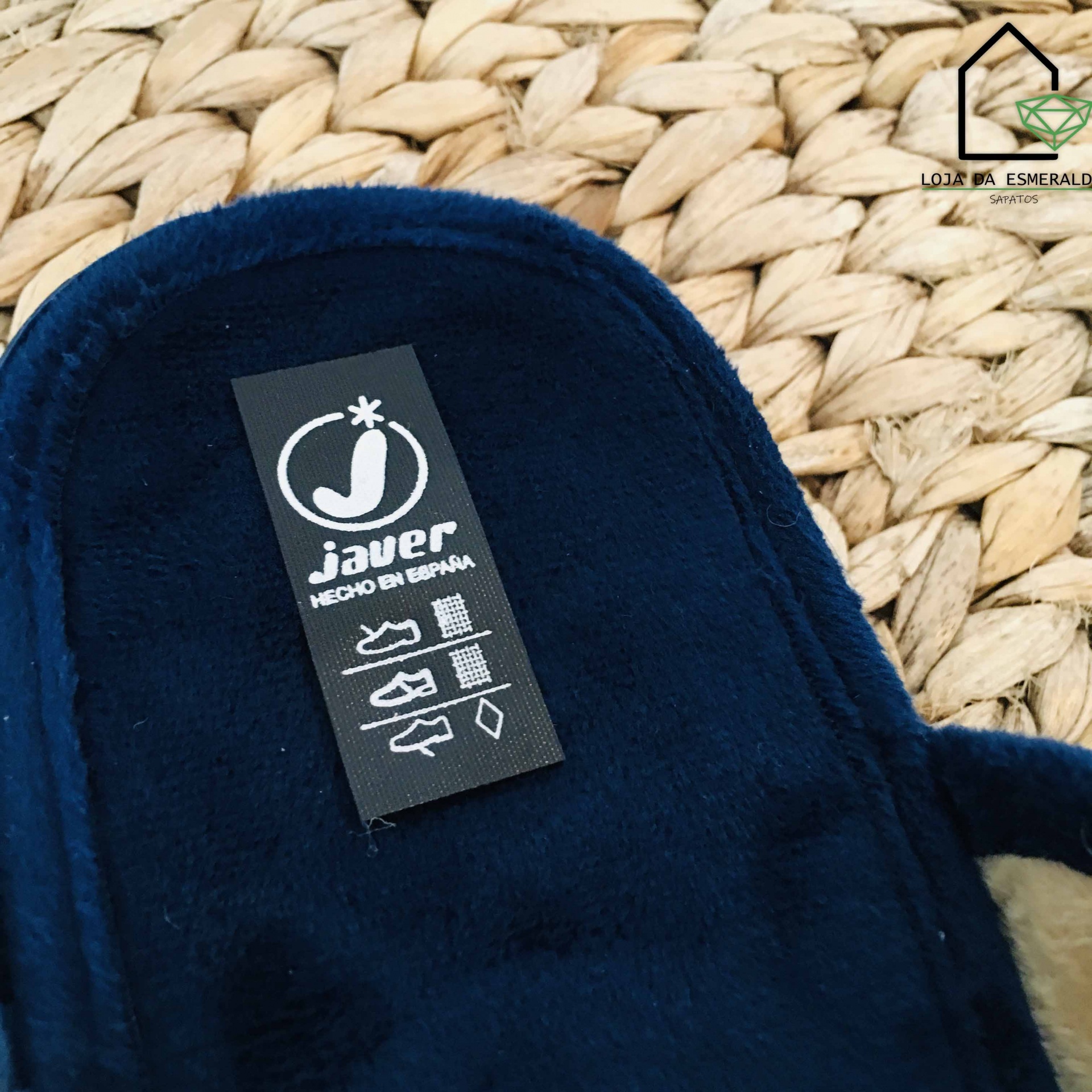 Etiqueta preta com texto 'javer HECHO EN ESPAÑA' numa pantufa azul escura aveludada sobre tecido trançado bege