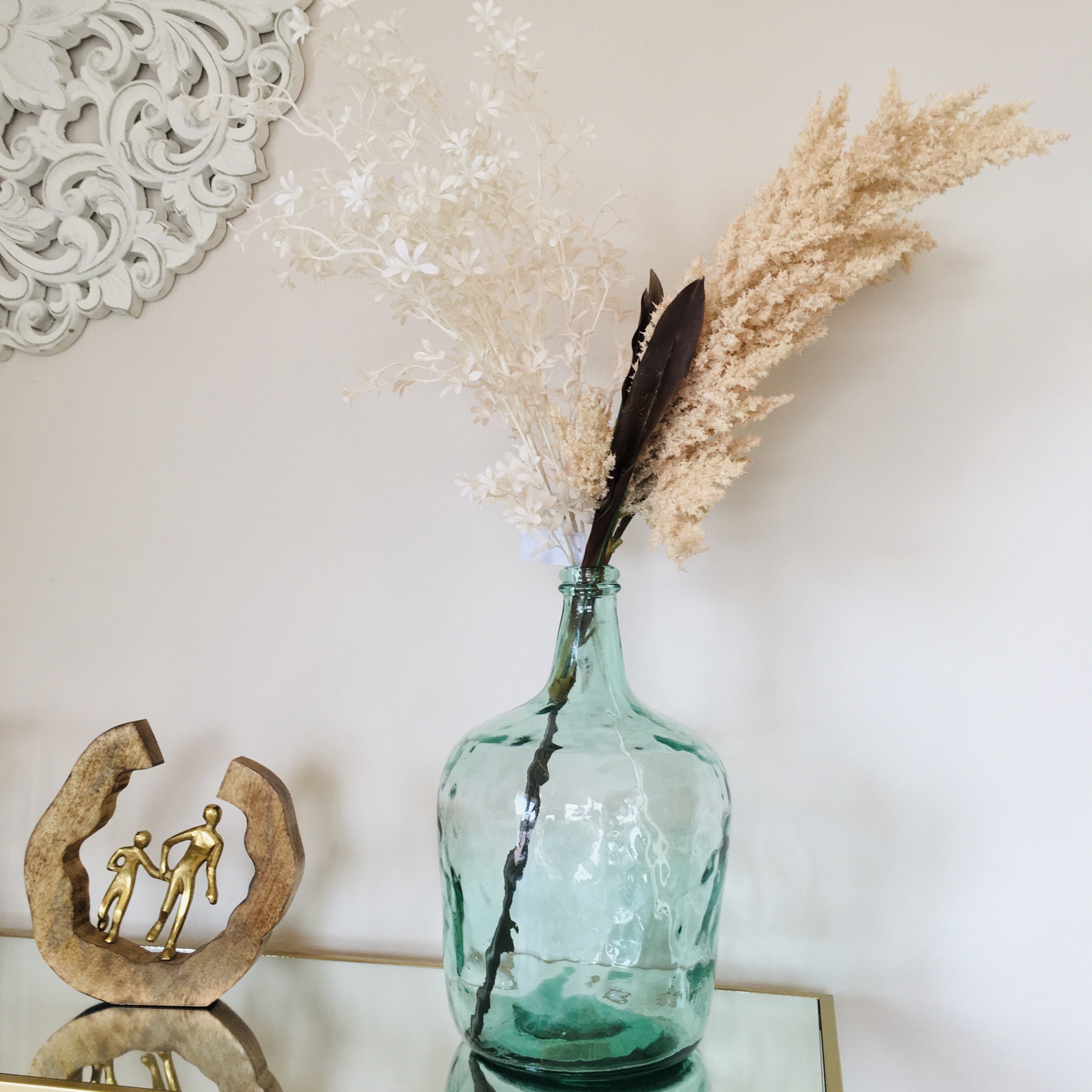 Garrafão de vidro verde com flores secas e escultura decorativa de figuras humanas em metal dourado sobre mesa espelhada