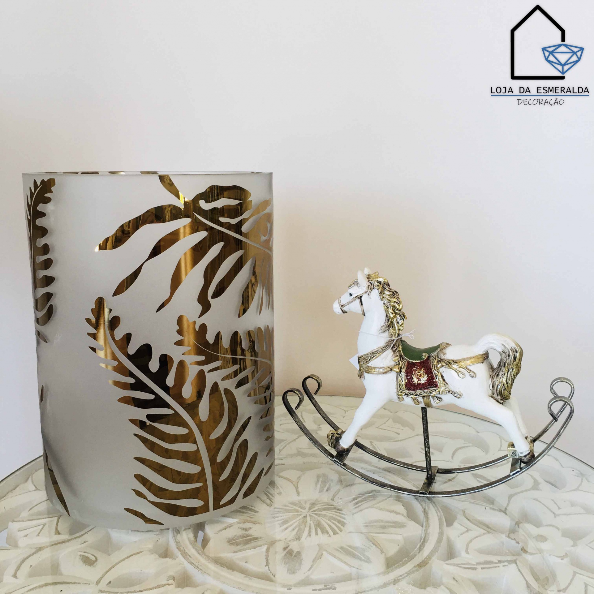 Castiçal de vidro fosco com folhas douradas ao lado de cavalo de balanço decorativo branco sobre superfície branca gravada.