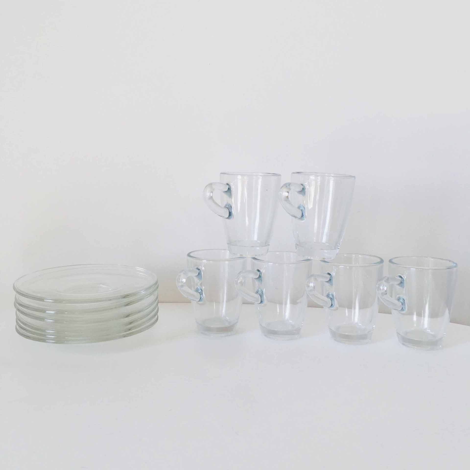 Seis chávenas de vidro com pegas em forma de coração e seis pequenos pratos de vidro empilhados numa superfície branca