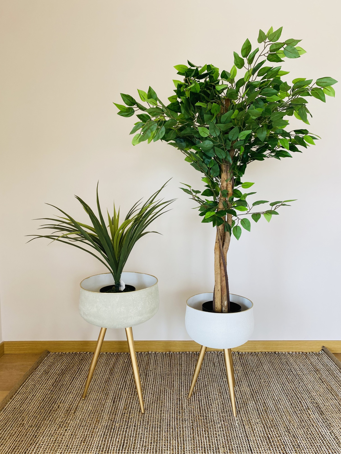 Dois vasos de plantas artificiais em suportes de madeira com plantas diferentes numa sala com parede creme e tapete castanho