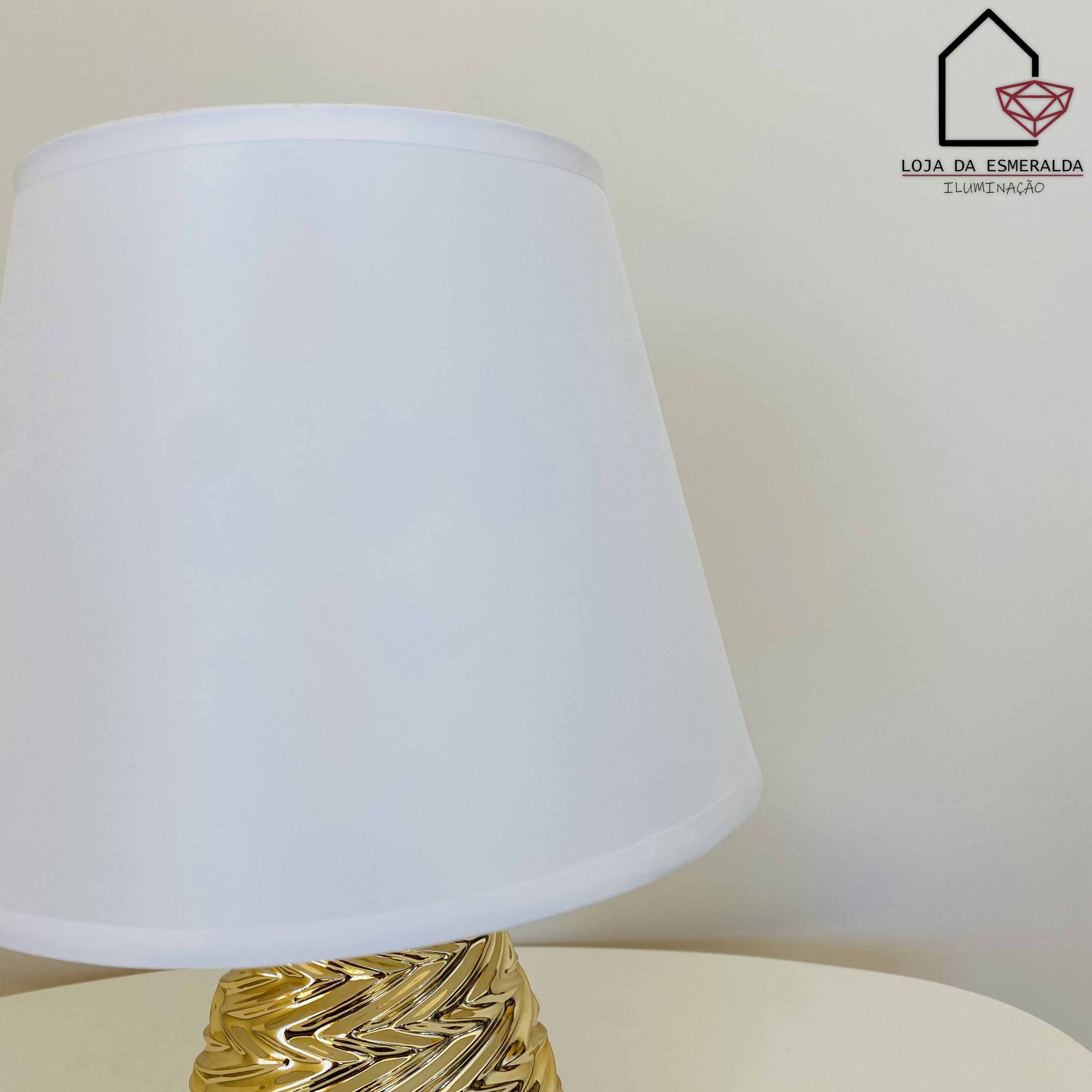 Candeeiro de mesa branco com base dourada texturada em fundo claro com logótipo