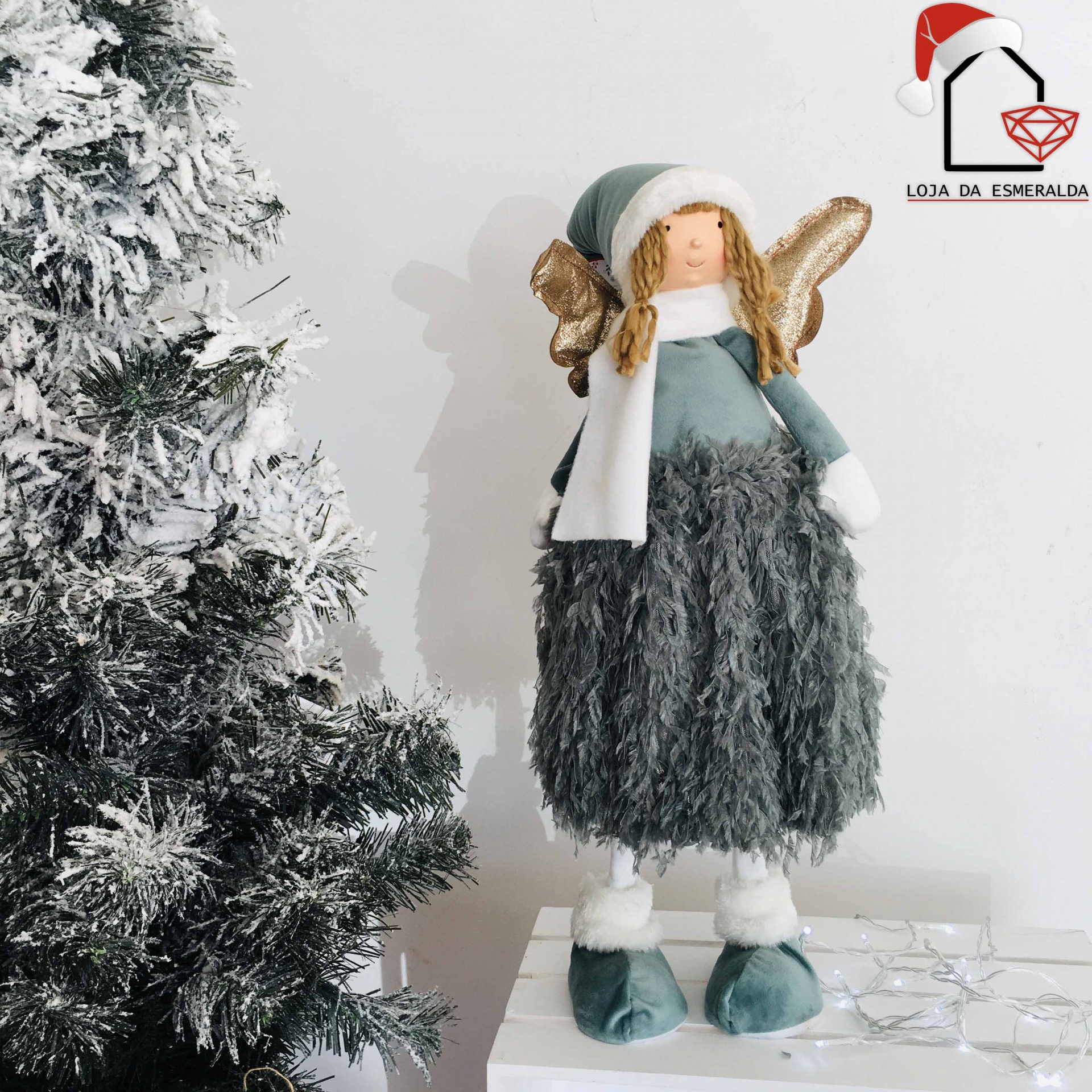 Anjo decorativo de Natal com vestido cinzento e azul, cachecol branco e asas douradas ao lado de árvore de Natal com neve