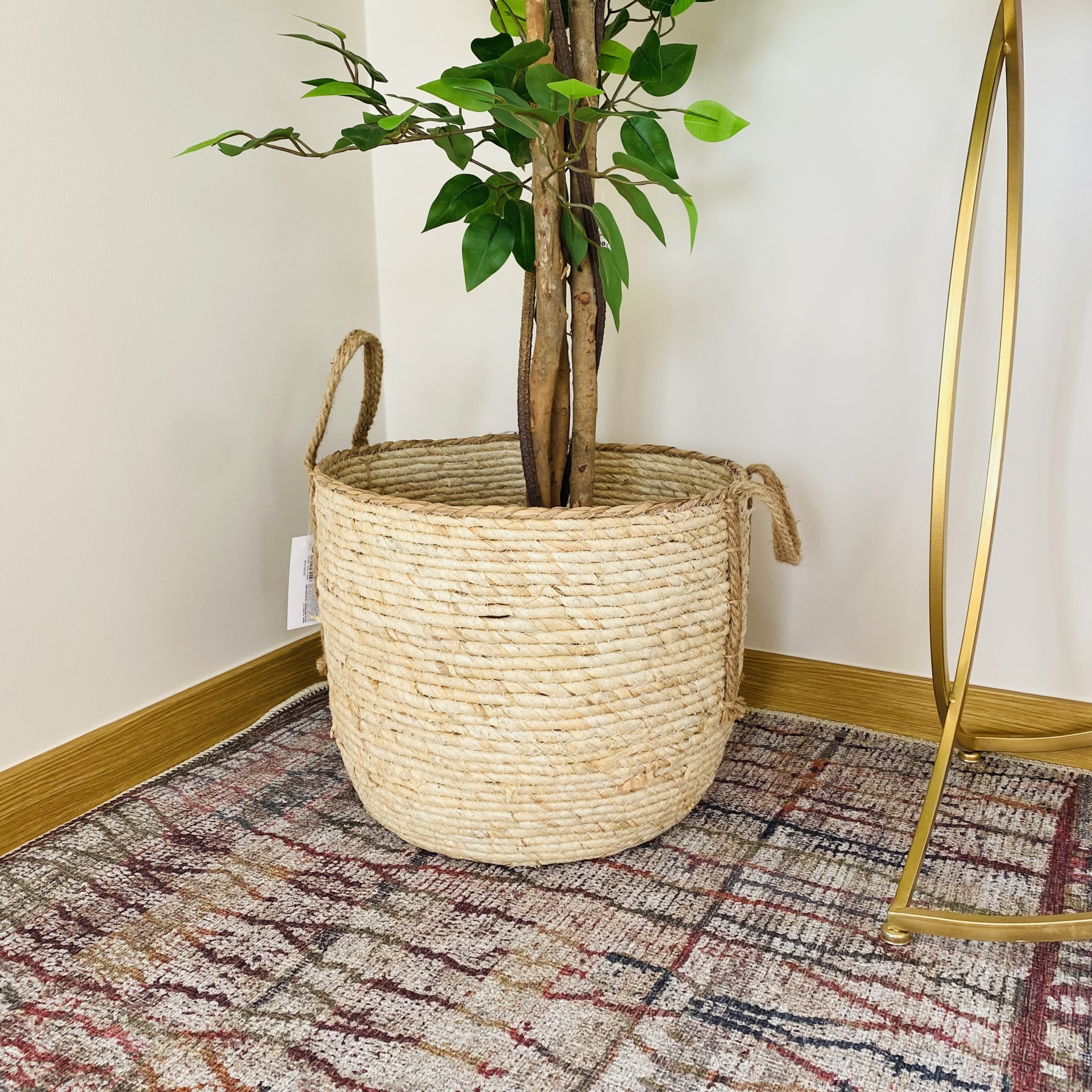 Cesto de corda natural com planta artificial em carpete colorida, perto de parede branca e rodapé madeira