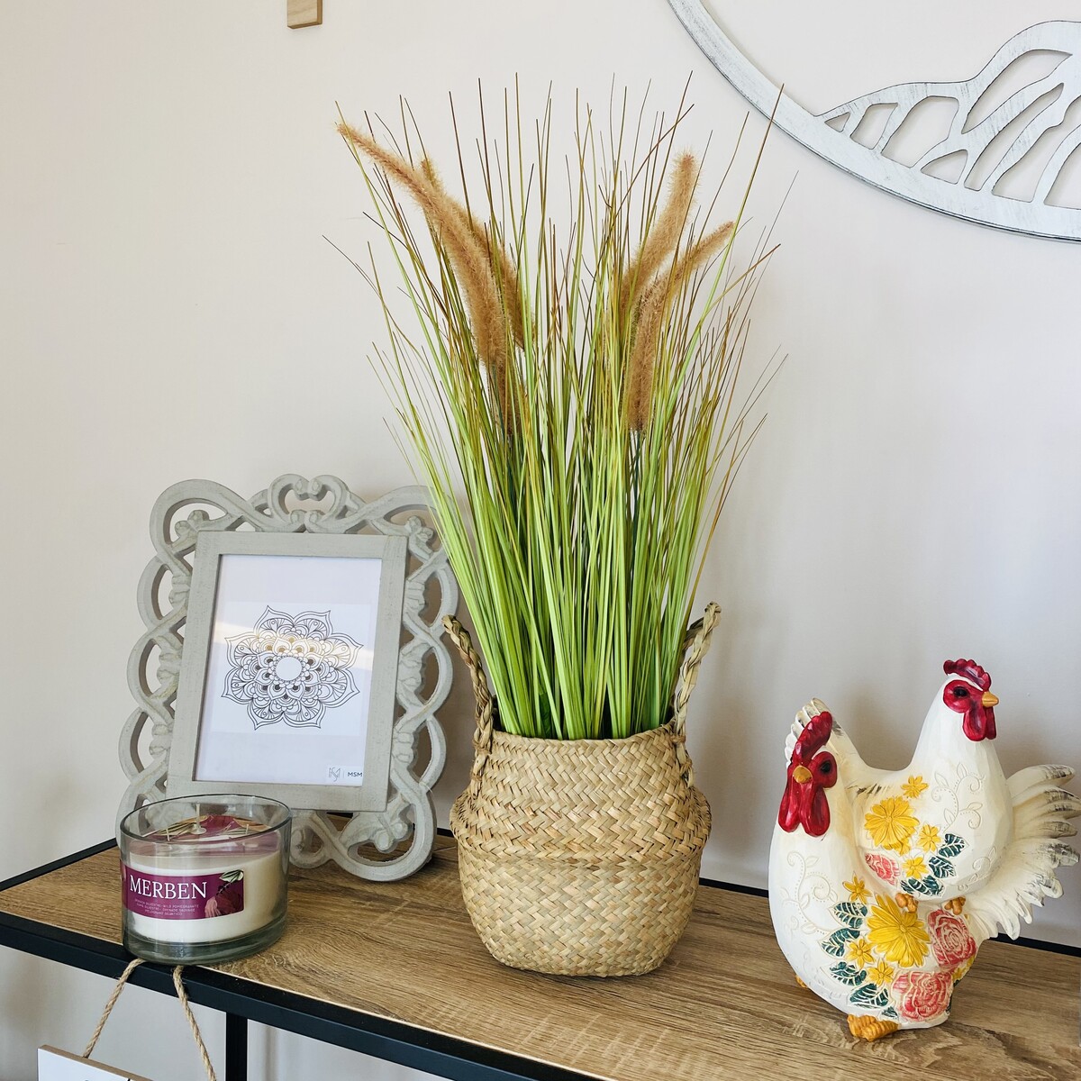 Vaso de cestos com planta artificial, moldura cinza e jarra decorativa de galo colorida numa mesa de madeira