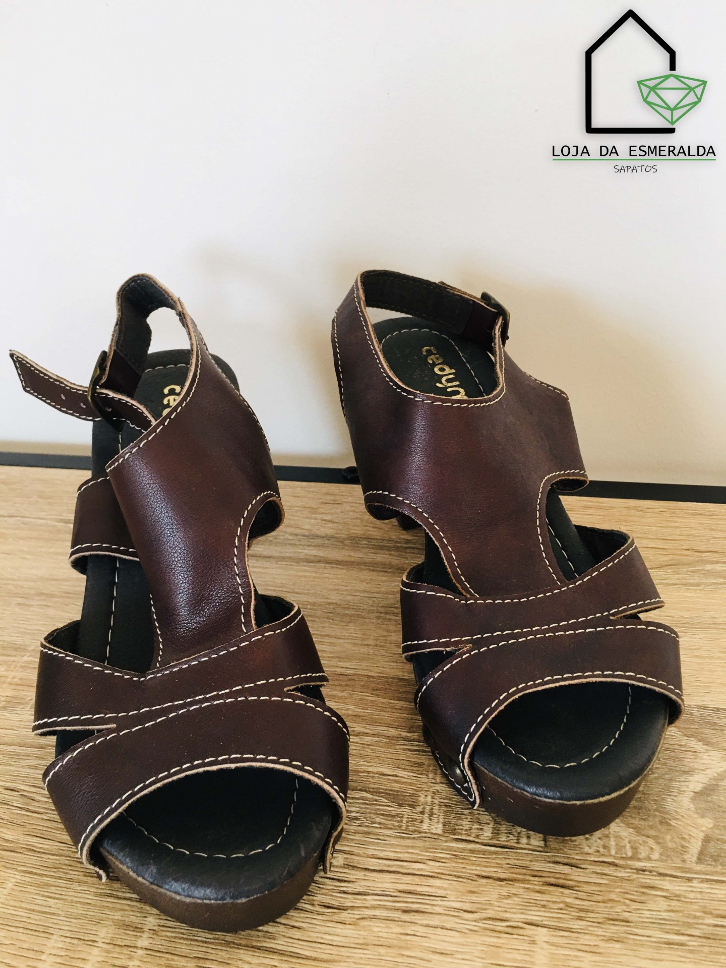 Sandálias castanhas em pele com costuras brancas sobre chão claro e logótipo da loja