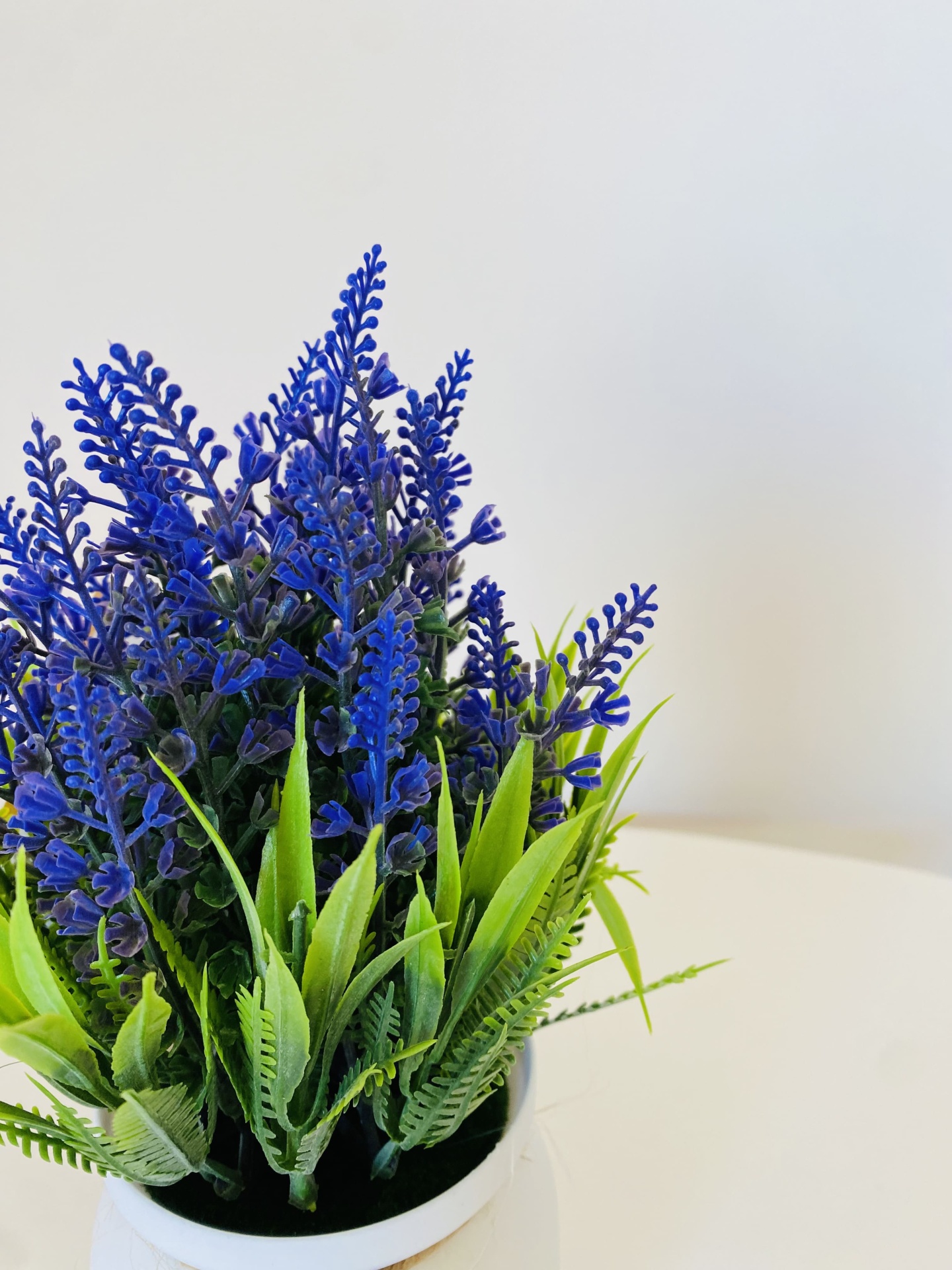 Arranjo floral artificial com flores azuis e folhas verdes em vaso branco sobre mesa branca