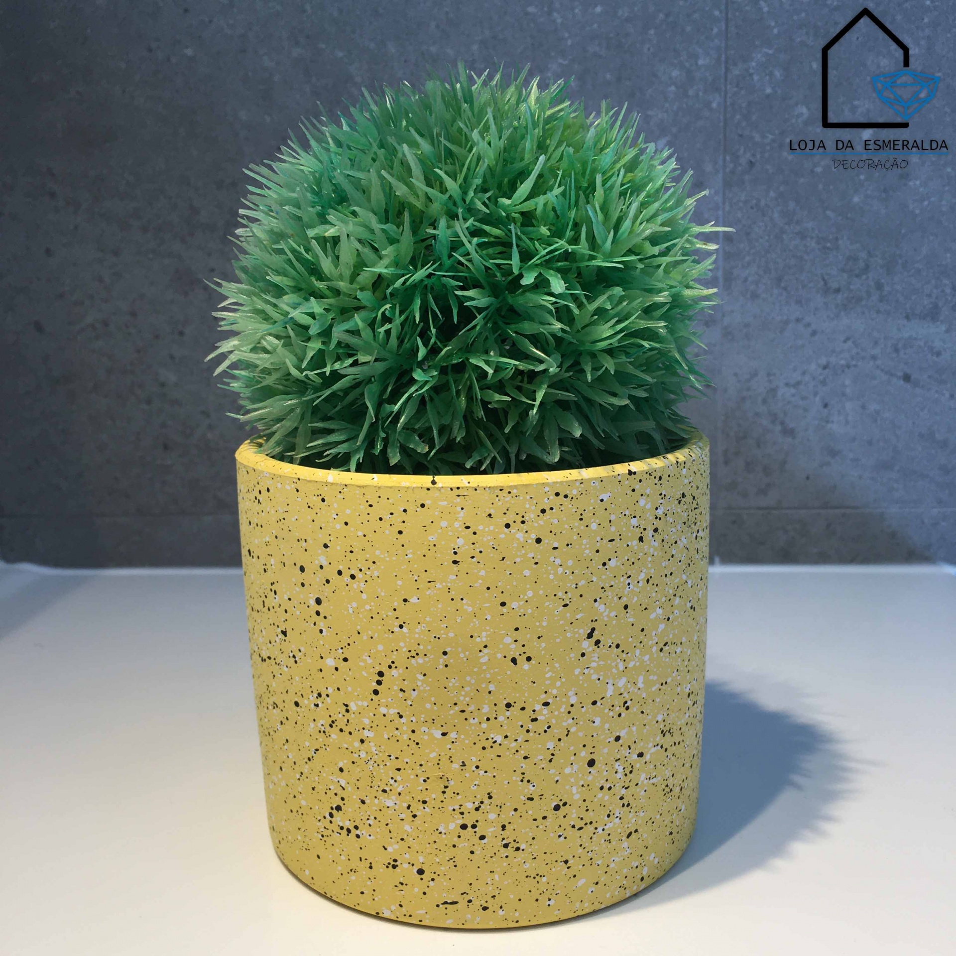 Vaso amarelo com planta artificial verde redonda em fundo cinza.