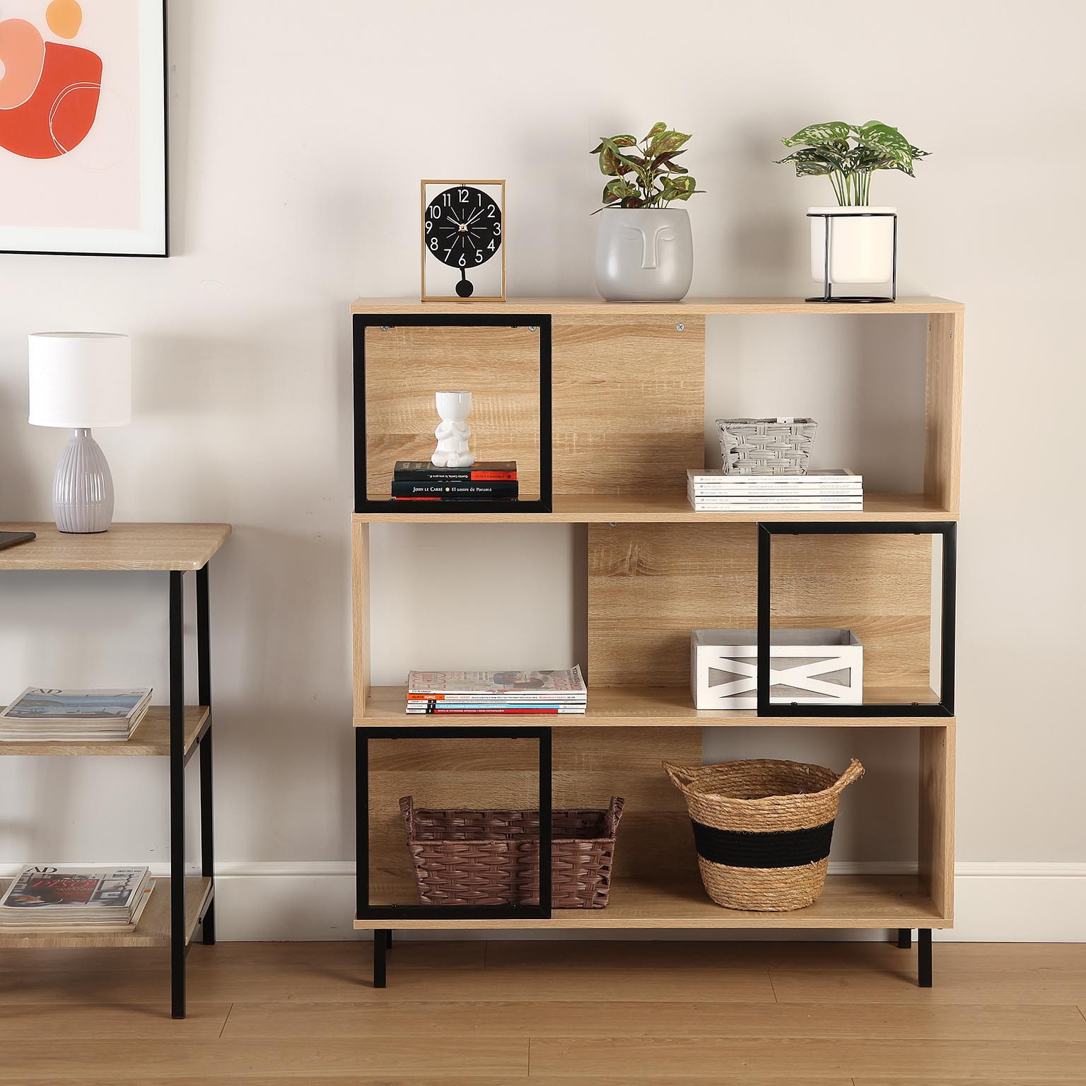 Estante de madeira clara com vasos, relógio, livros e cestos, junto a parede branca com quadro e mesa lateral