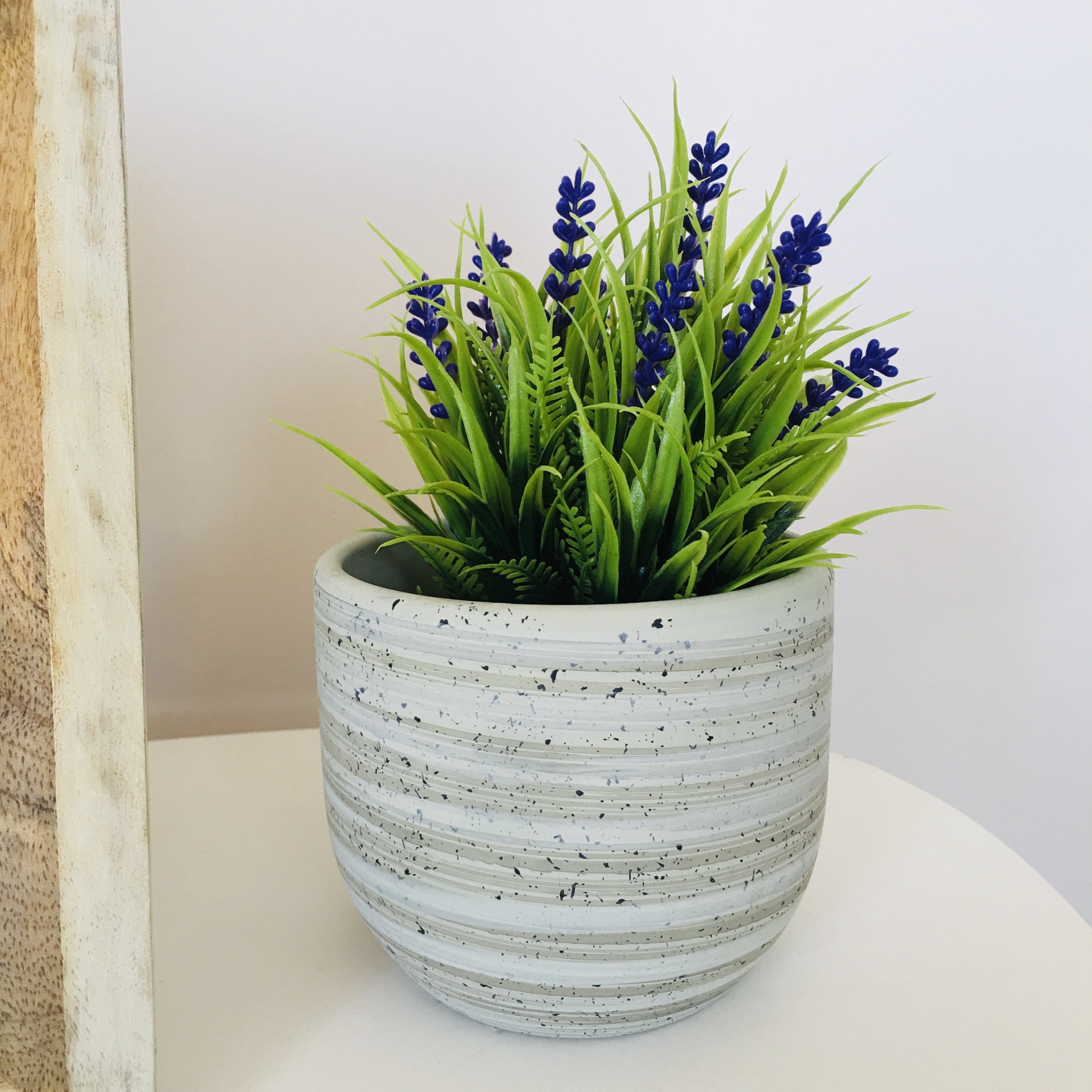 Vaso cinzento com planta artificial verde e flores roxas em cima de mesa branca
