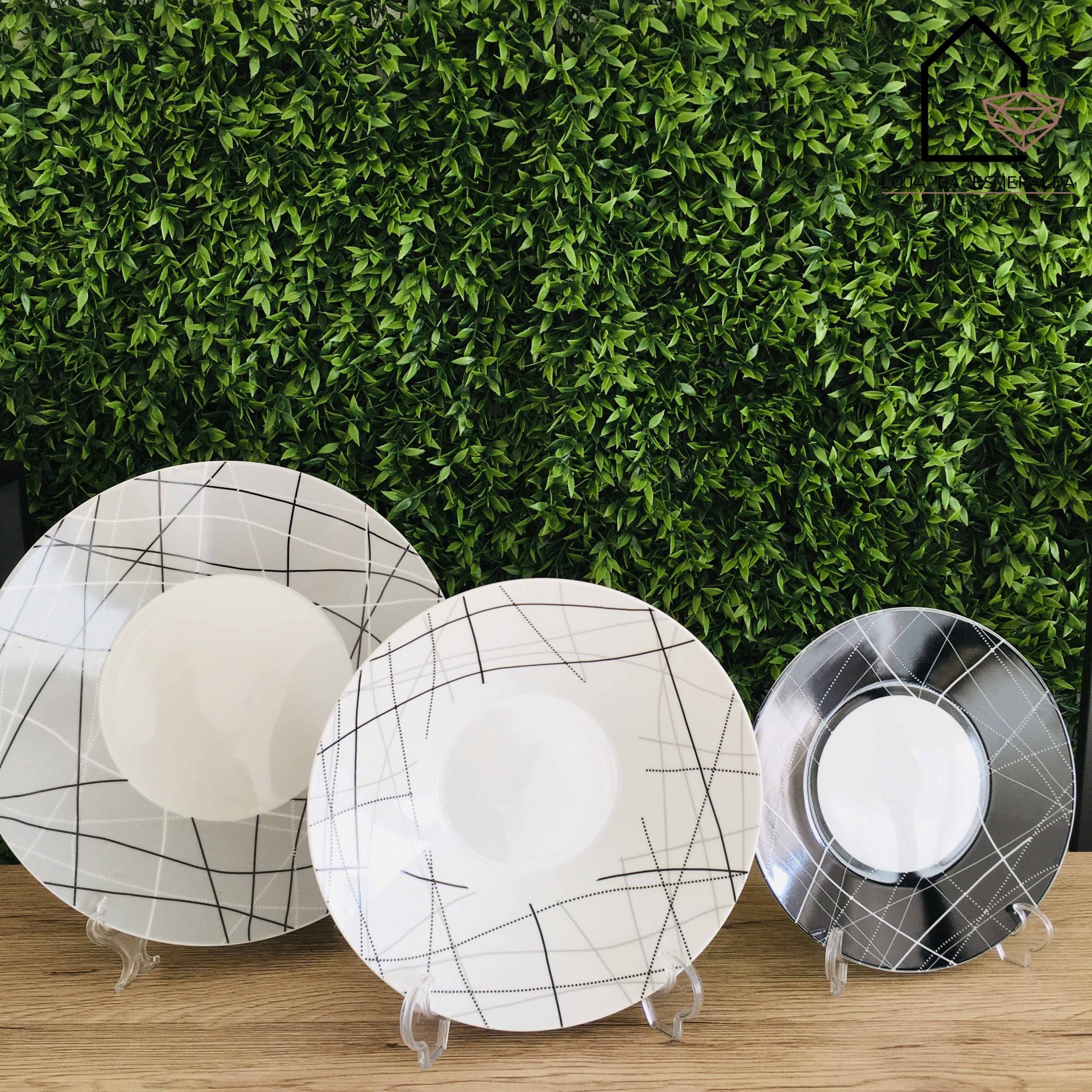 Três pratos cerâmicos com padrão de linhas pretas sobre fundo branco e cinza, expostos com suporte transparente frente a uma parede verde