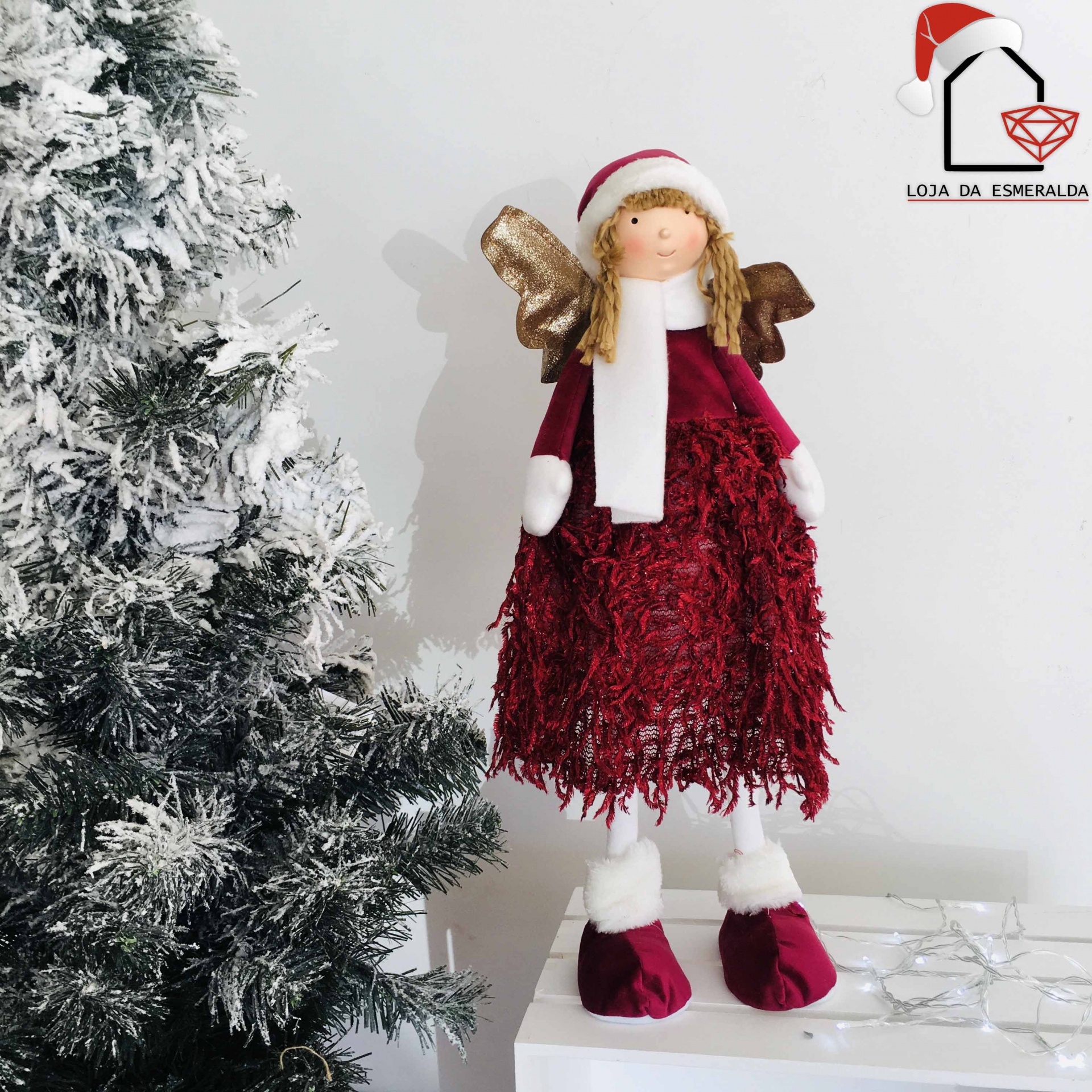 Anjo decorativo de Natal com vestido vermelho e asas douradas ao lado de árvore de Natal
