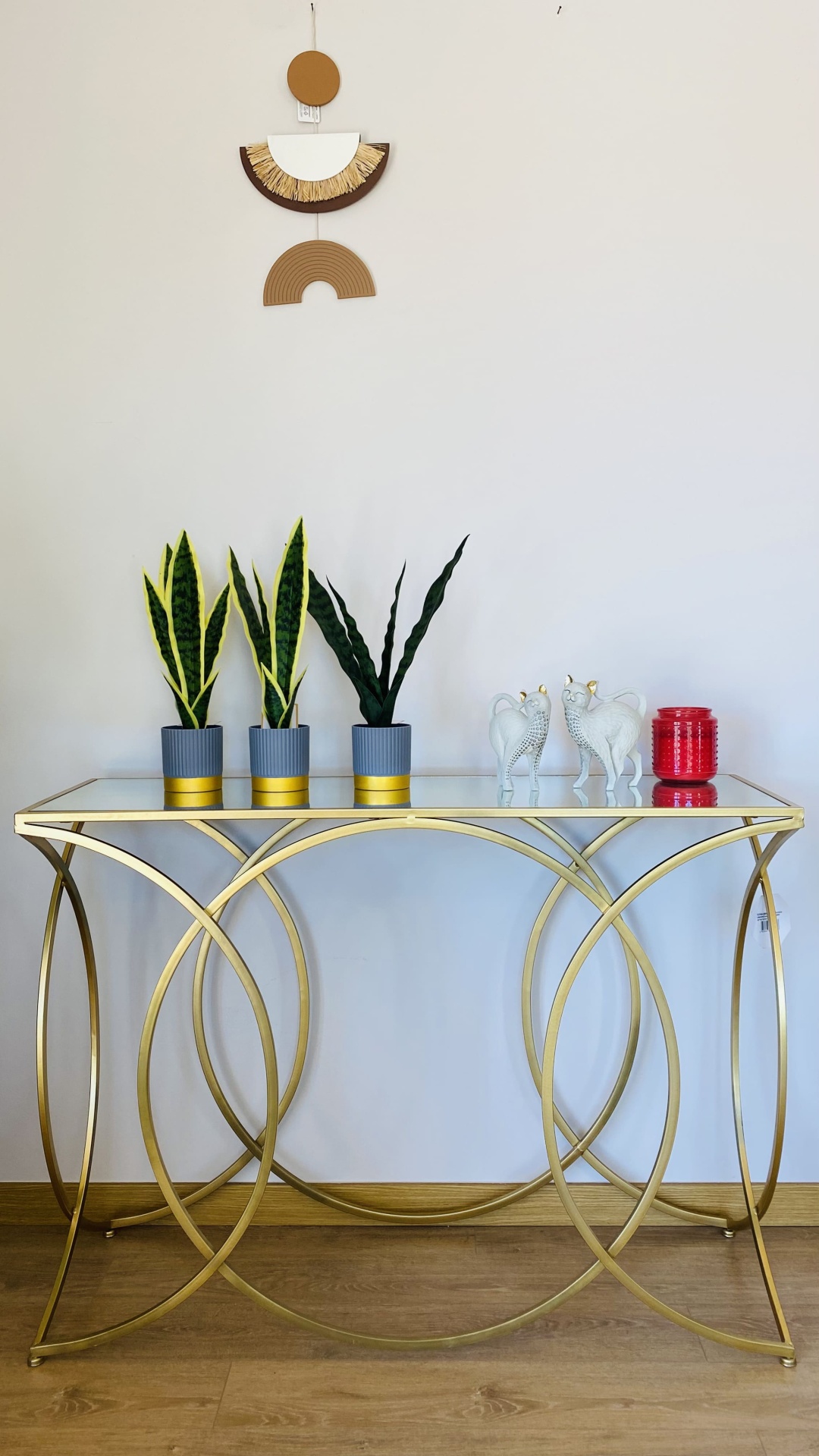 Mesa dourada com vidro com plantas, estátuas de gatos e vaso vermelho em sala com parede branca e decoração pendurada.