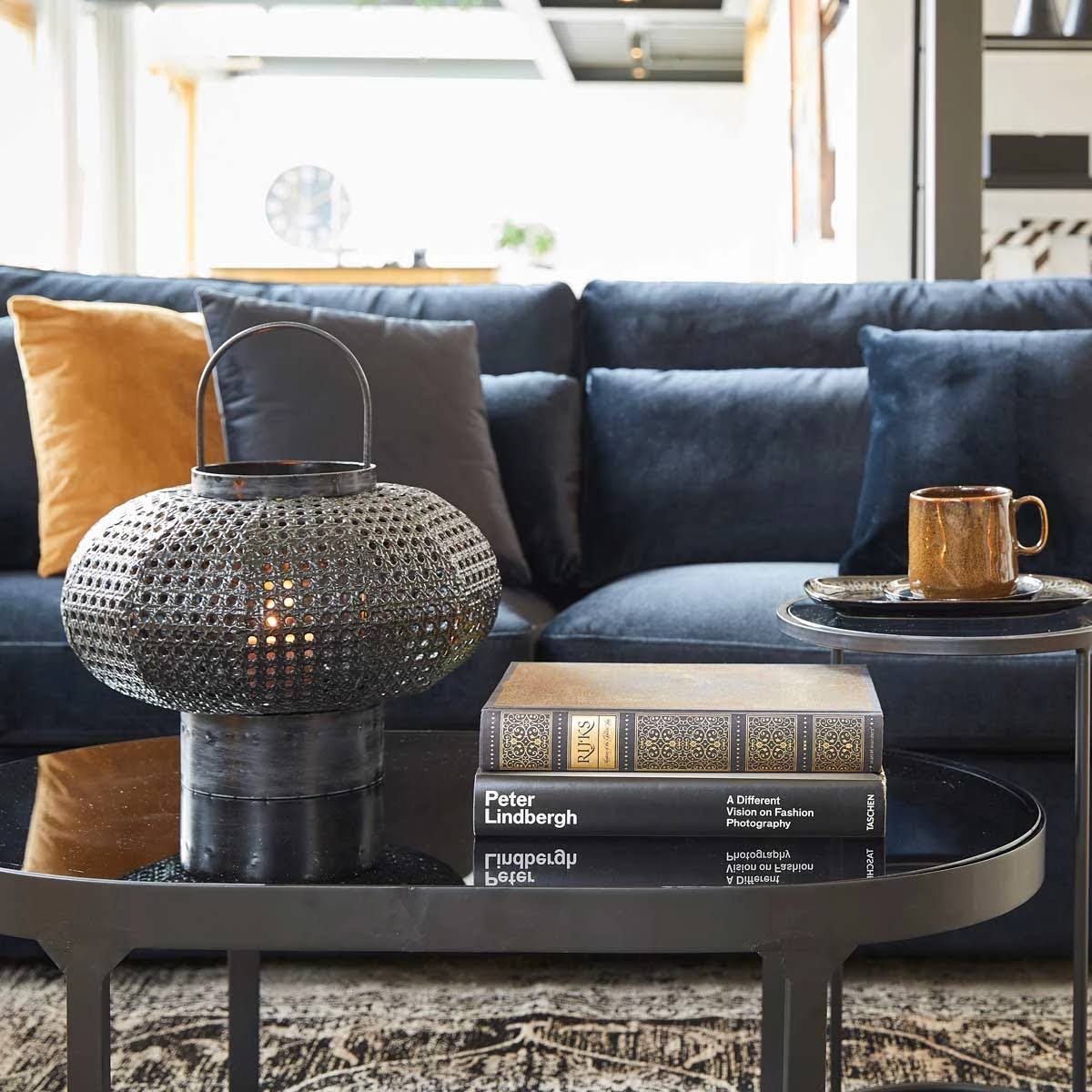 Mesa de centro com lanterna metálica preta, livros empilhados e caneca castanha, sofá azul ao fundo