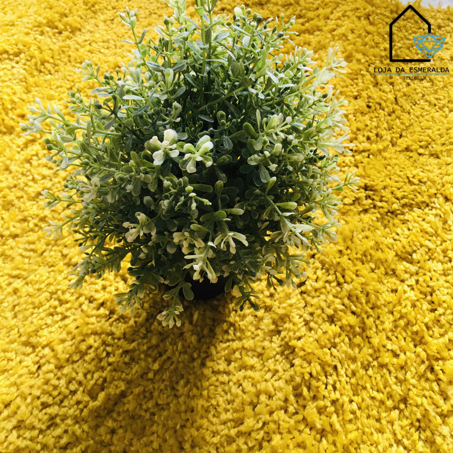 Vaso preto com planta artificial verde e flores brancas sobre tapete amarelo felpudo com logotipo CASA DA ESMERALDA