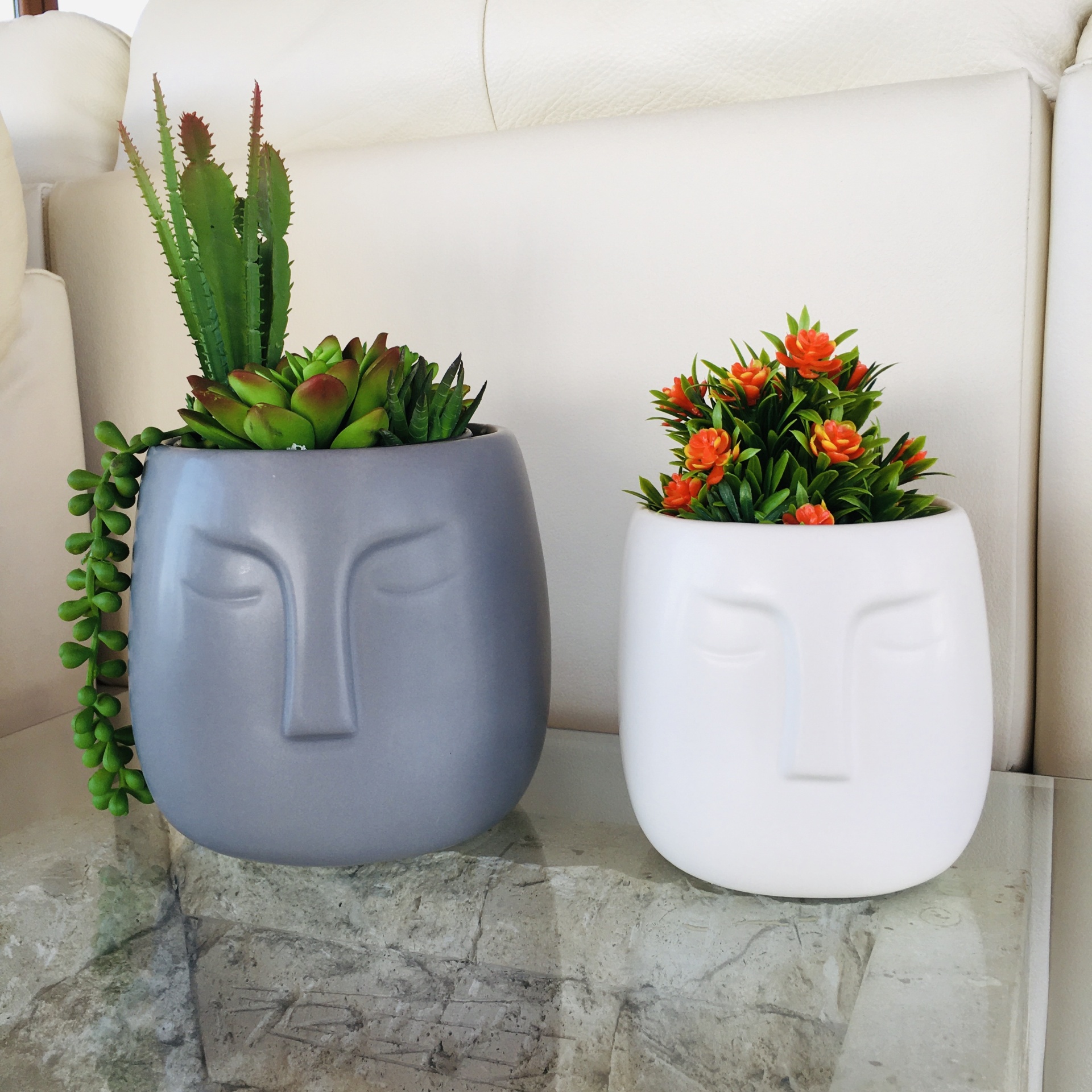 Dois vasos cerâmicos com formato de rosto e plantas, um cinza com suculentas e outro branco com flores laranja.