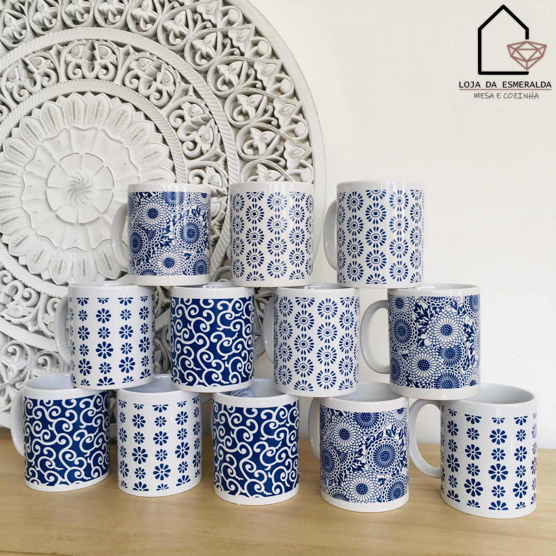 Canecas brancas com padrões azuis em vários designs, dispostas em pirâmide sobre uma mesa de madeira, com decoração branca ao fundo.
