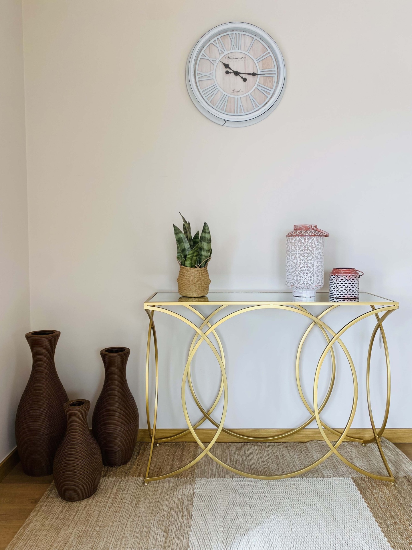 Relógio branco de parede, mesa dourada com vidro, vasos castanhos, planta e lanternas decorativas num ambiente interior.