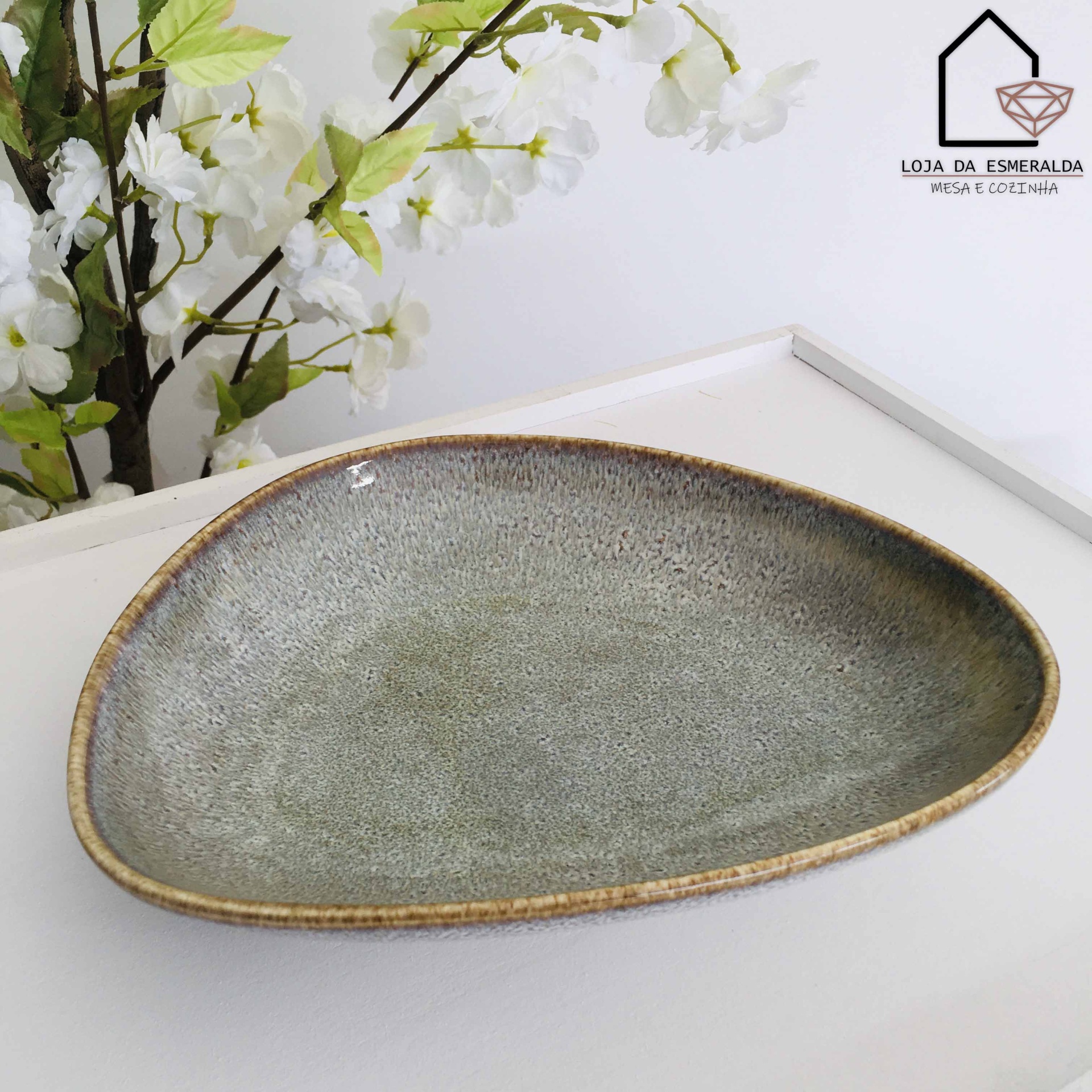 Prato triangular cinza esverdeado com borda castanha e flores brancas ao fundo