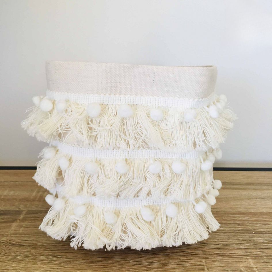 Cesto decorativo em tecido cru com franjas e pompons brancos sobre superfície de madeira