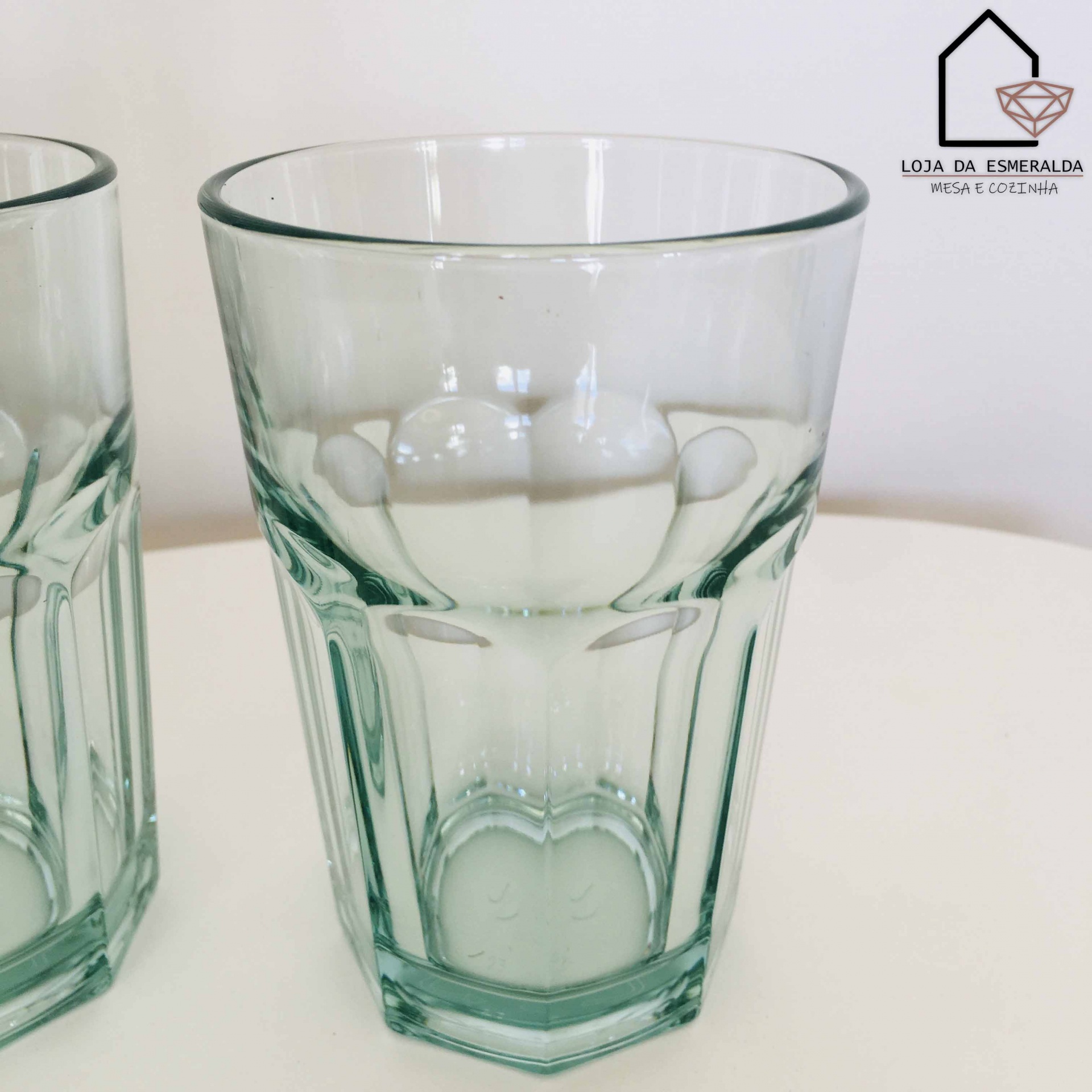Copo de vidro transparente com padrão facetado e logotipo Loja da Esmeralda no fundo