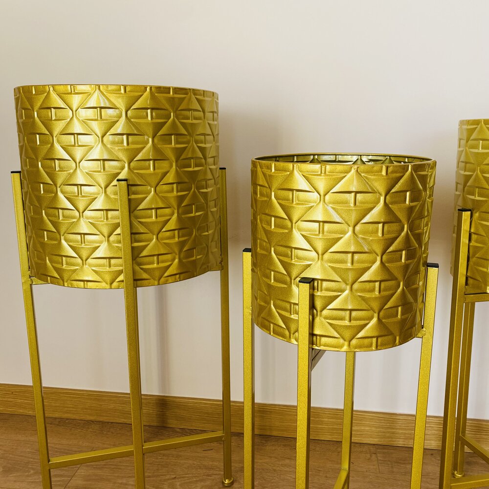 Três cachepots dourados com suporte metálico em chão de madeira