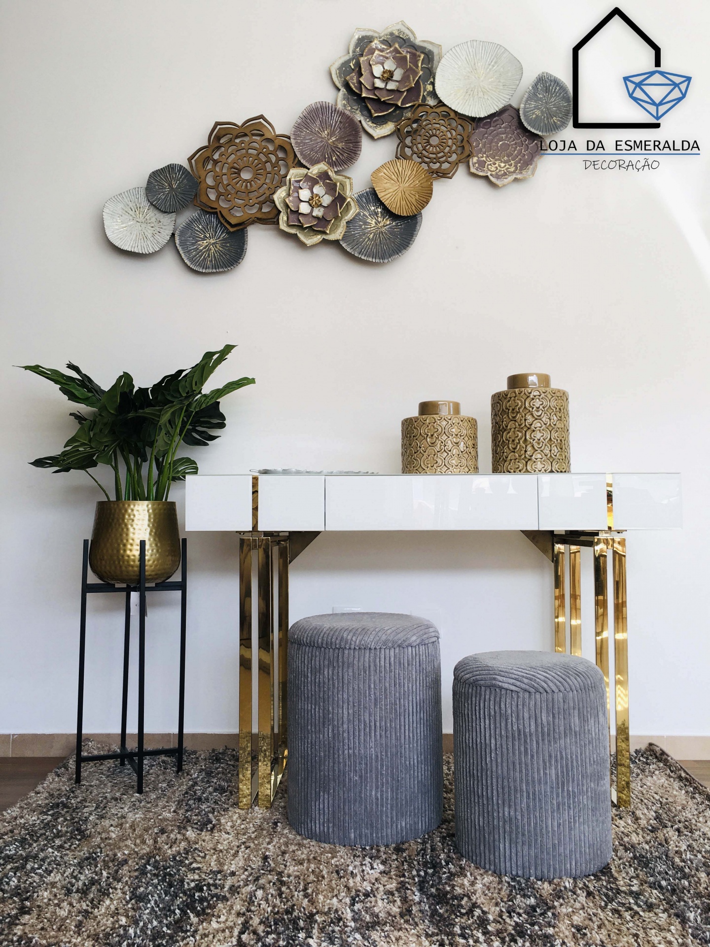 Conjunto de móveis e decoração com aparador branco, dois pufes cinza, vaso dourado com planta e painel floral na parede
