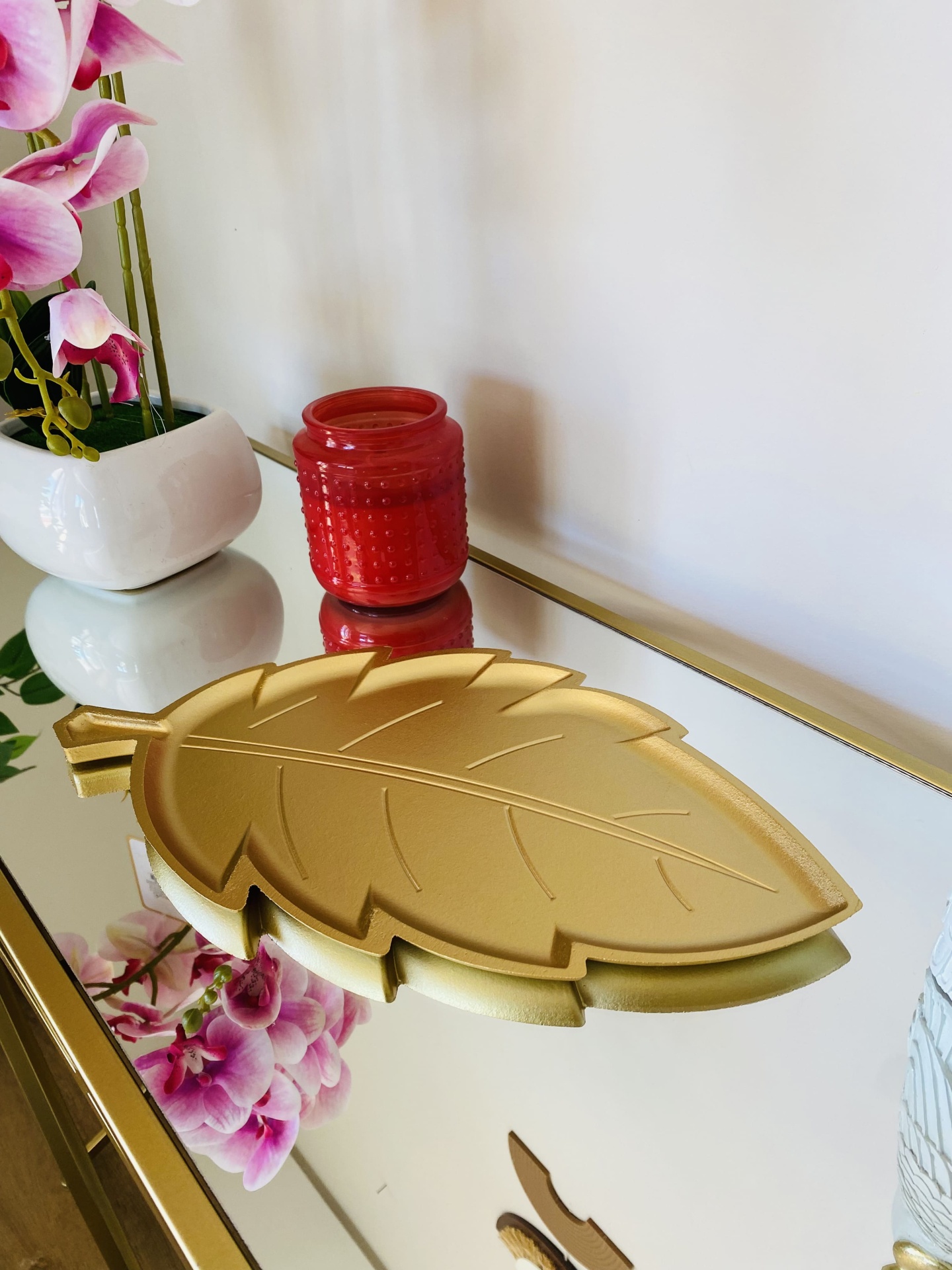 Placa dourada em forma de folha numa superfície espelhada com vaso de flores cor-de-rosa e vela vermelha