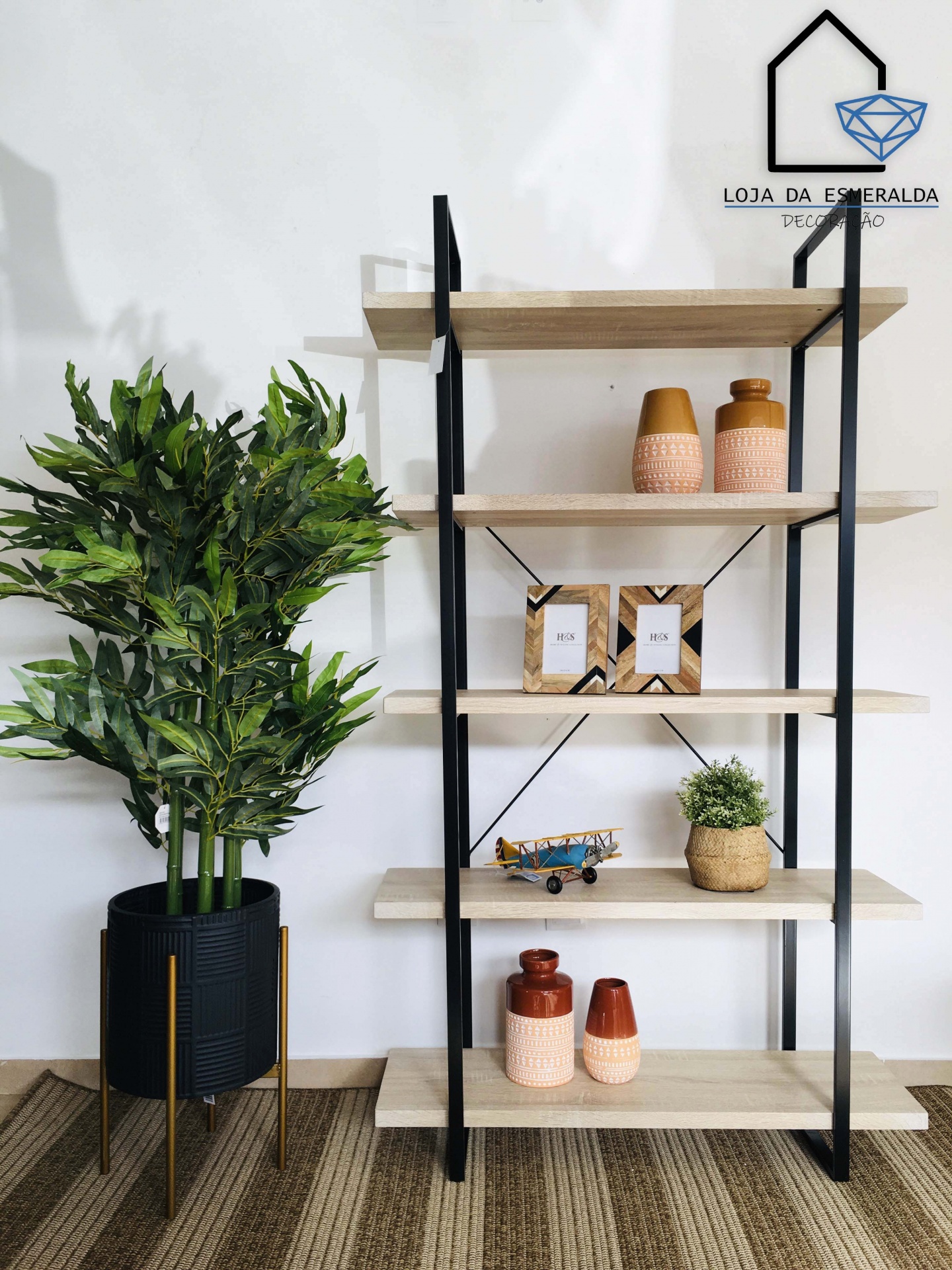 Estante preta e madeira clara com vasos e decoração, planta verde ao lado em suporte metálico dourado