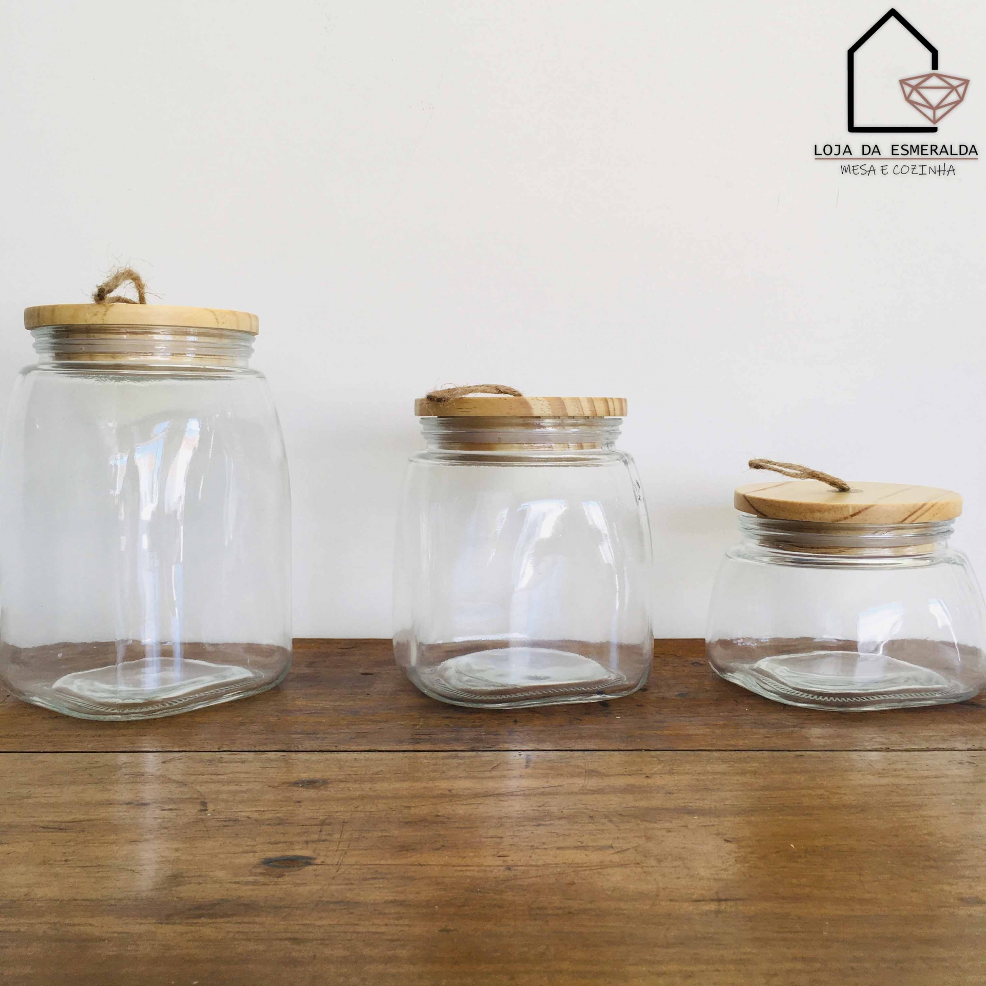 Três frascos de vidro com tampas de madeira em diferentes tamanhos sobre uma mesa de madeira.