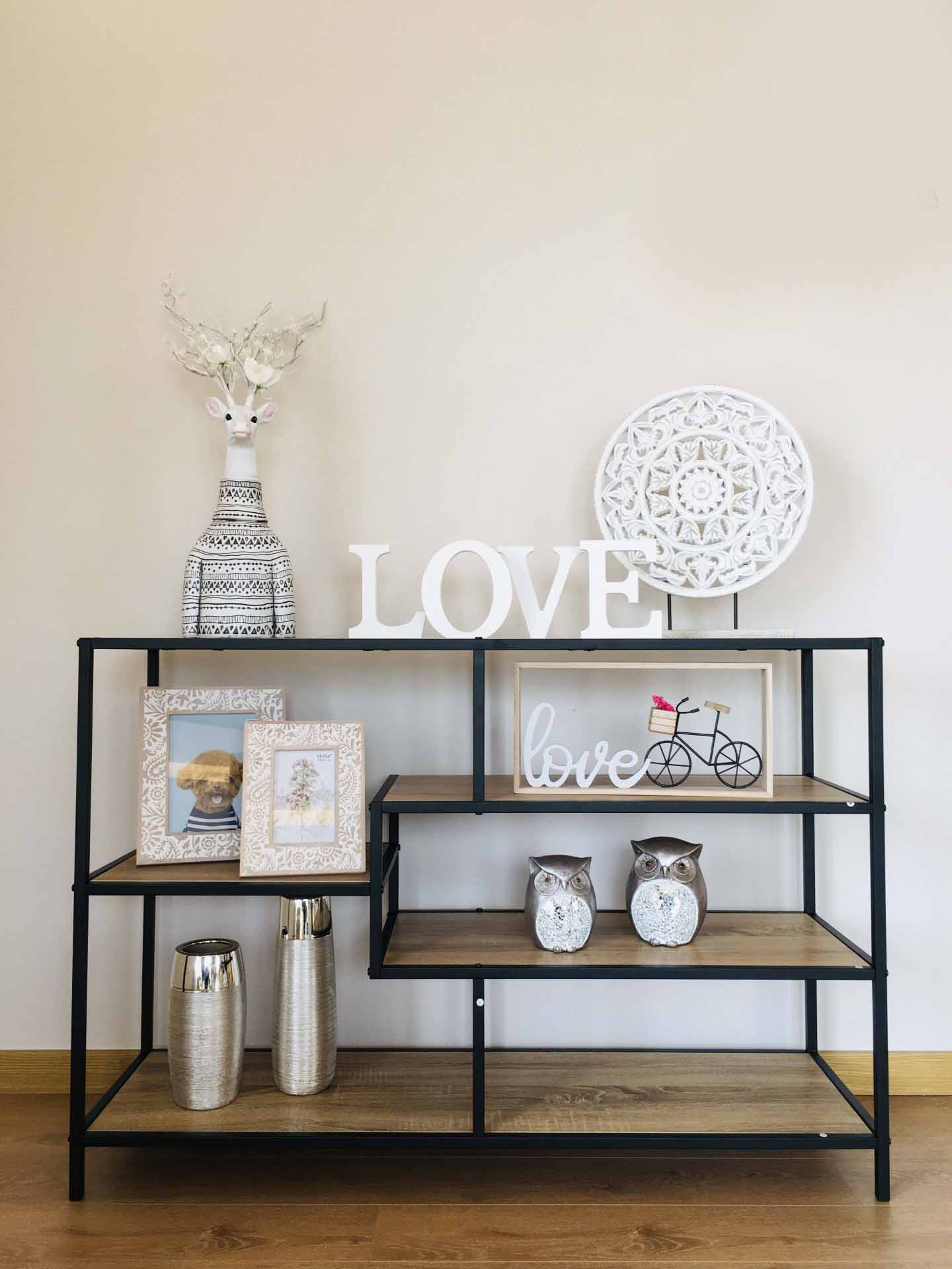 Estante preta com prateleiras de madeira e objetos decorativos incluindo cabeça de veado, palavra LOVE, prato branco, molduras, corujas e vasos prateados.