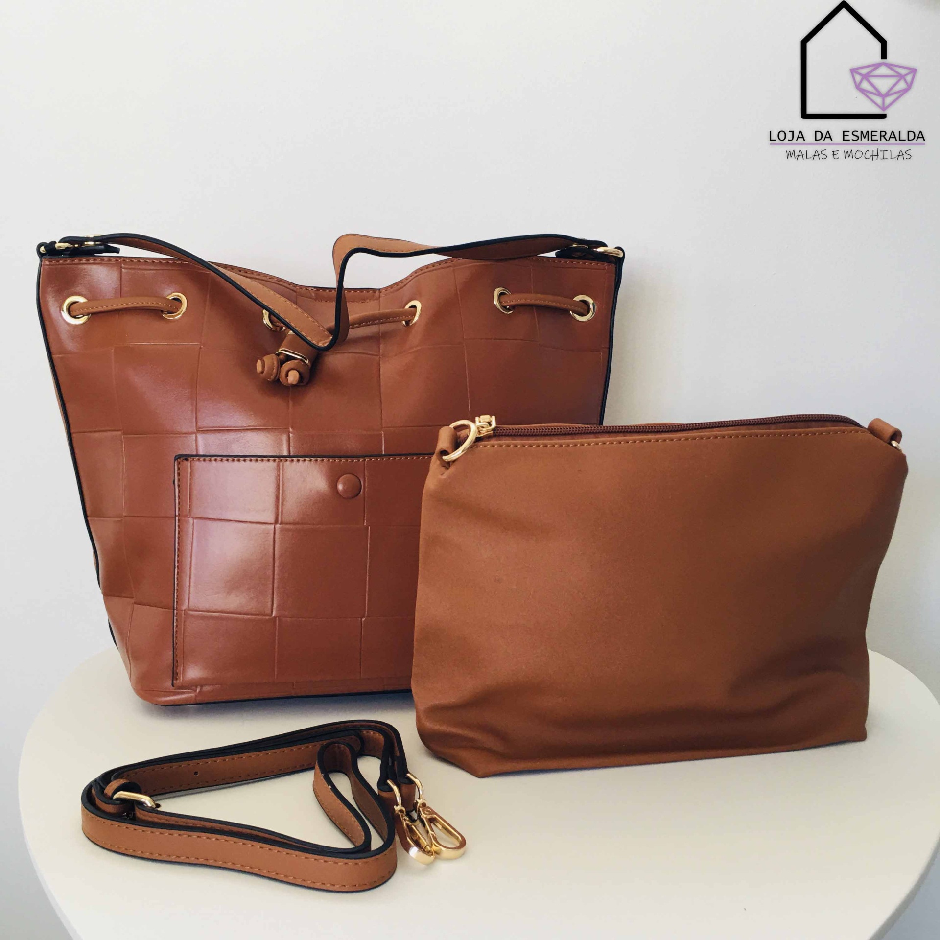 Conjunto de duas bolsas castanhas, uma grande com padrão quadrado e outra pequena lisa, com alça destacável, sobre mesa branca e logo da loja no fundo.