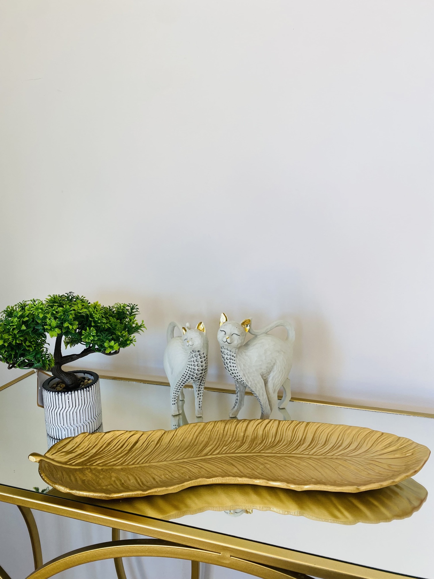 Decoração com placa dourada em forma de folha, vaso com planta artificial e esculturas de gatos cinzentos com detalhes dourados.