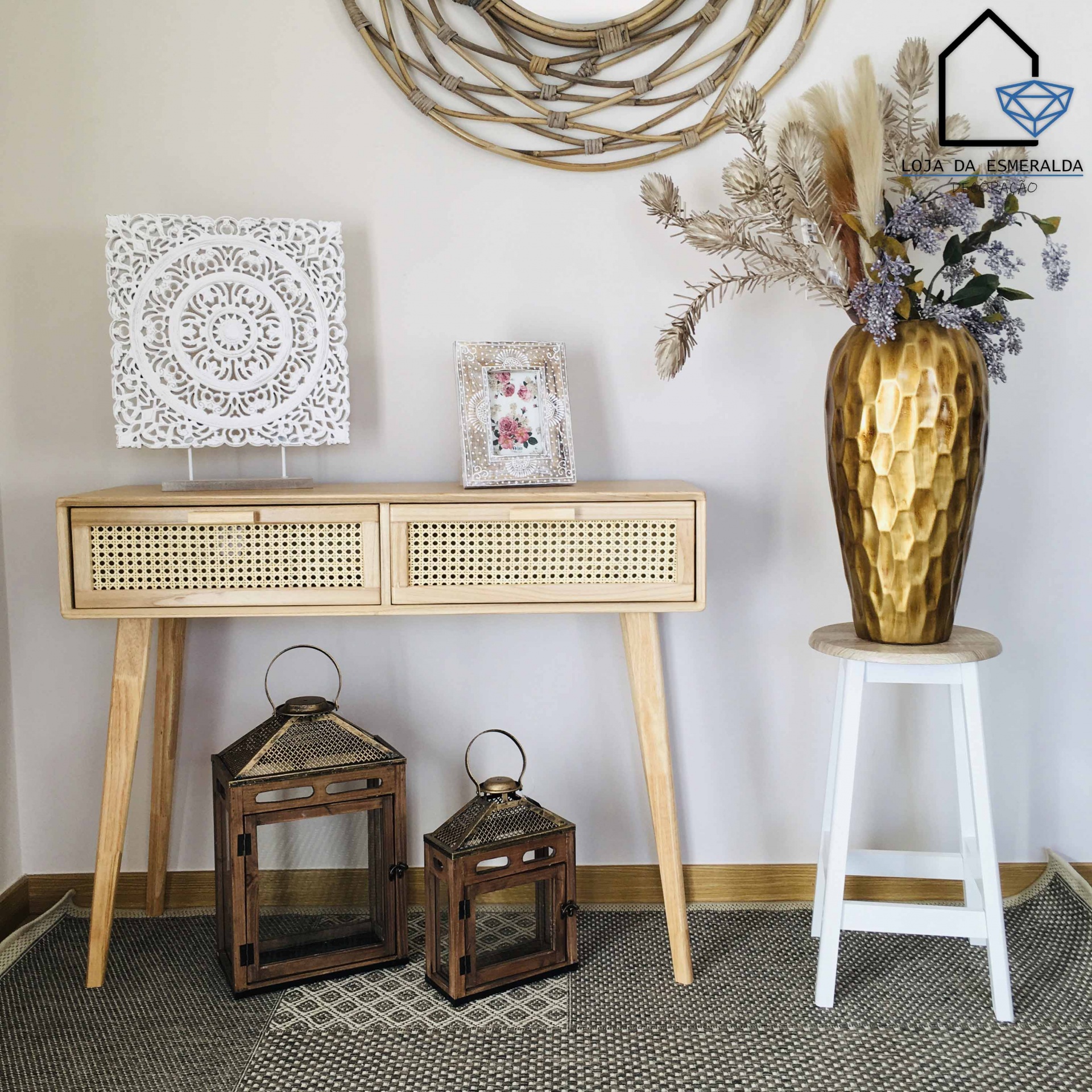 Mesa de madeira clara com gavetas de rattan, lanternas, vaso dourado com flores secas, banco branco e decoração de parede clara
