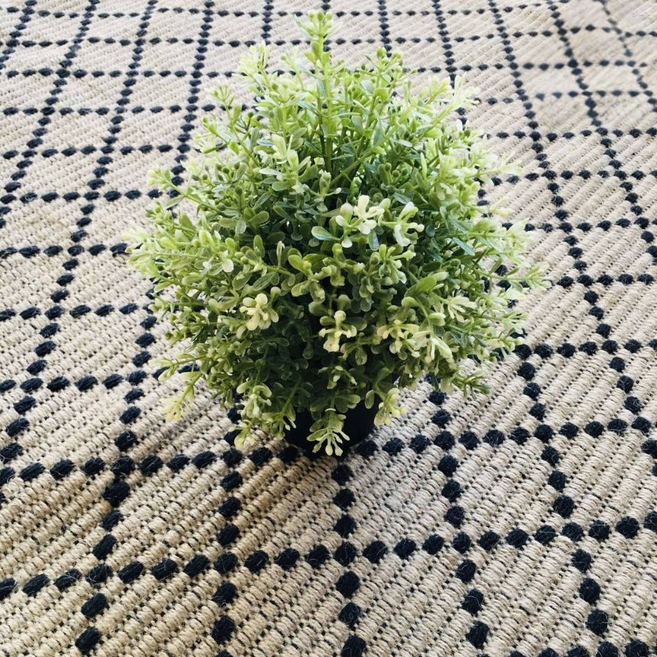 Planta artificial verde em vaso preto sobre tapete bege com padrão preto