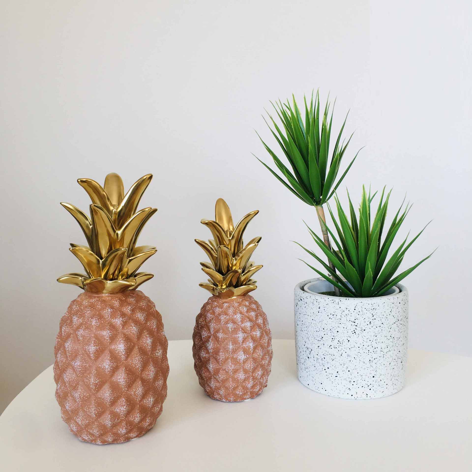 Duas figuras decorativas de ananás castanho com topo dourado e vaso branco com planta artificial verde