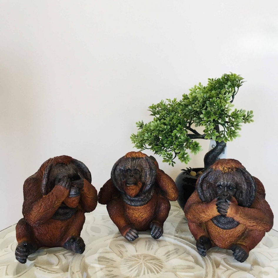 Três estatuetas de macacos castanhos em mesa com planta artificial ao fundo