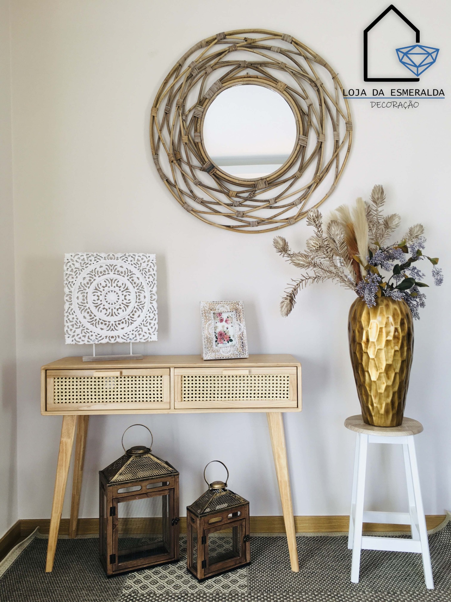 Móvel em madeira clara com gavetas de palhinha, espelho trançado redondo, vaso dourado com flores secas e dois candeeiros de madeira escura