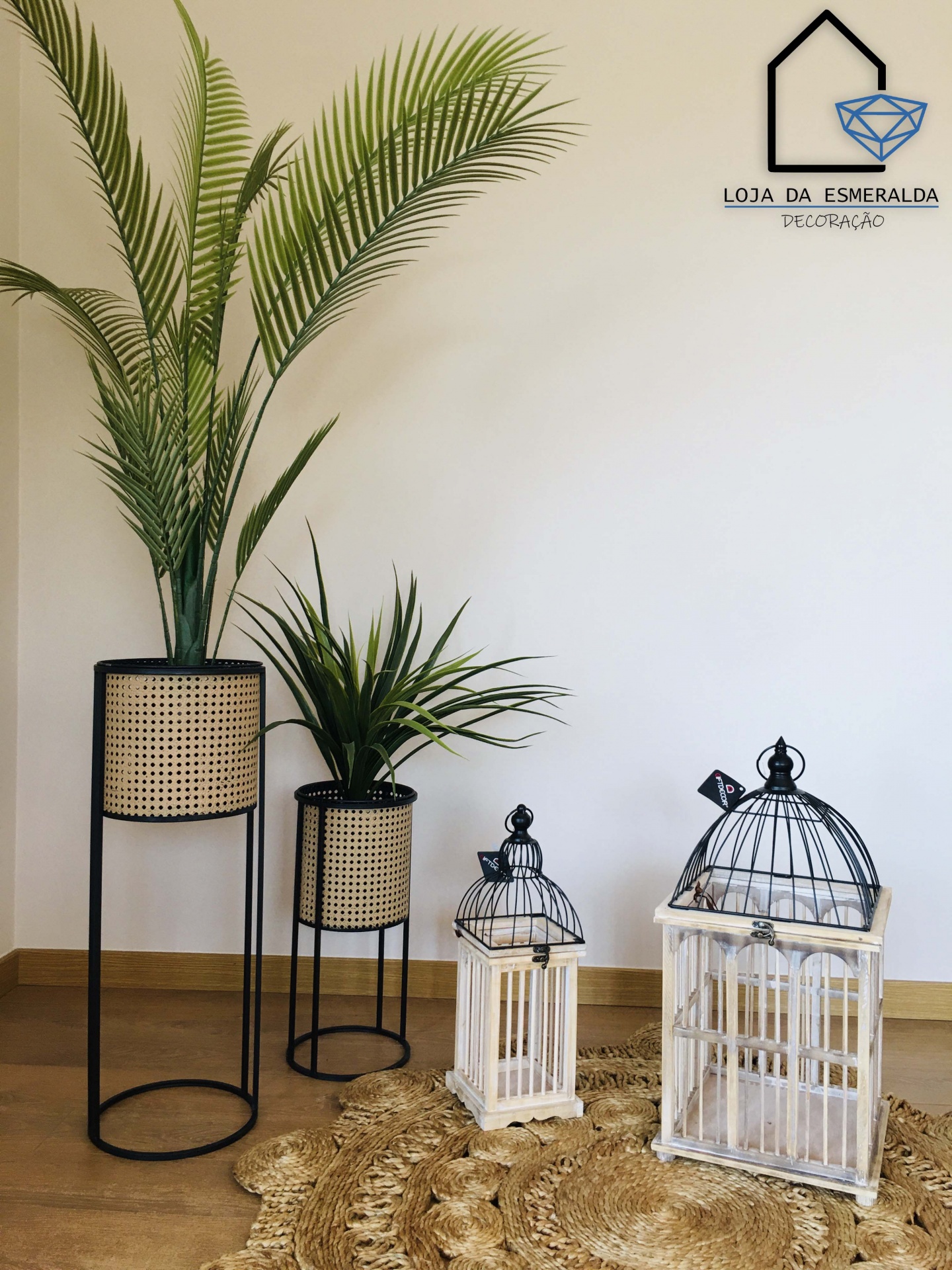 Dois vasos com plantas artificiais em suportes de metal preto e cestos de vime, duas lanternas brancas e tapete de palha num ambiente interior