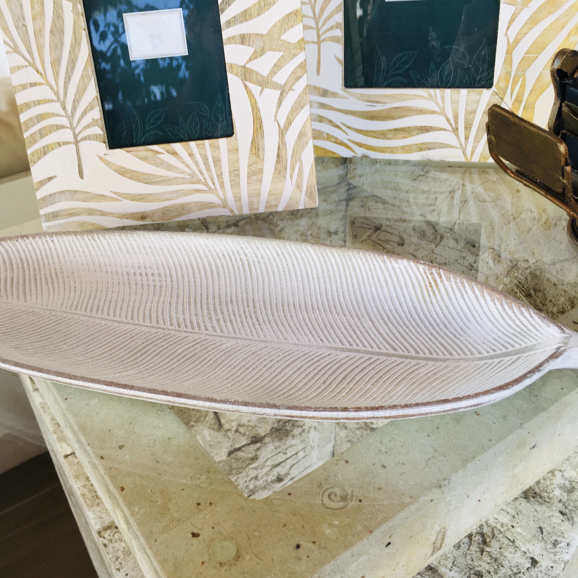 Prato decorativo em forma de folha bege claro com textura, sobre mesa de pedra com molduras de fotos ao fundo