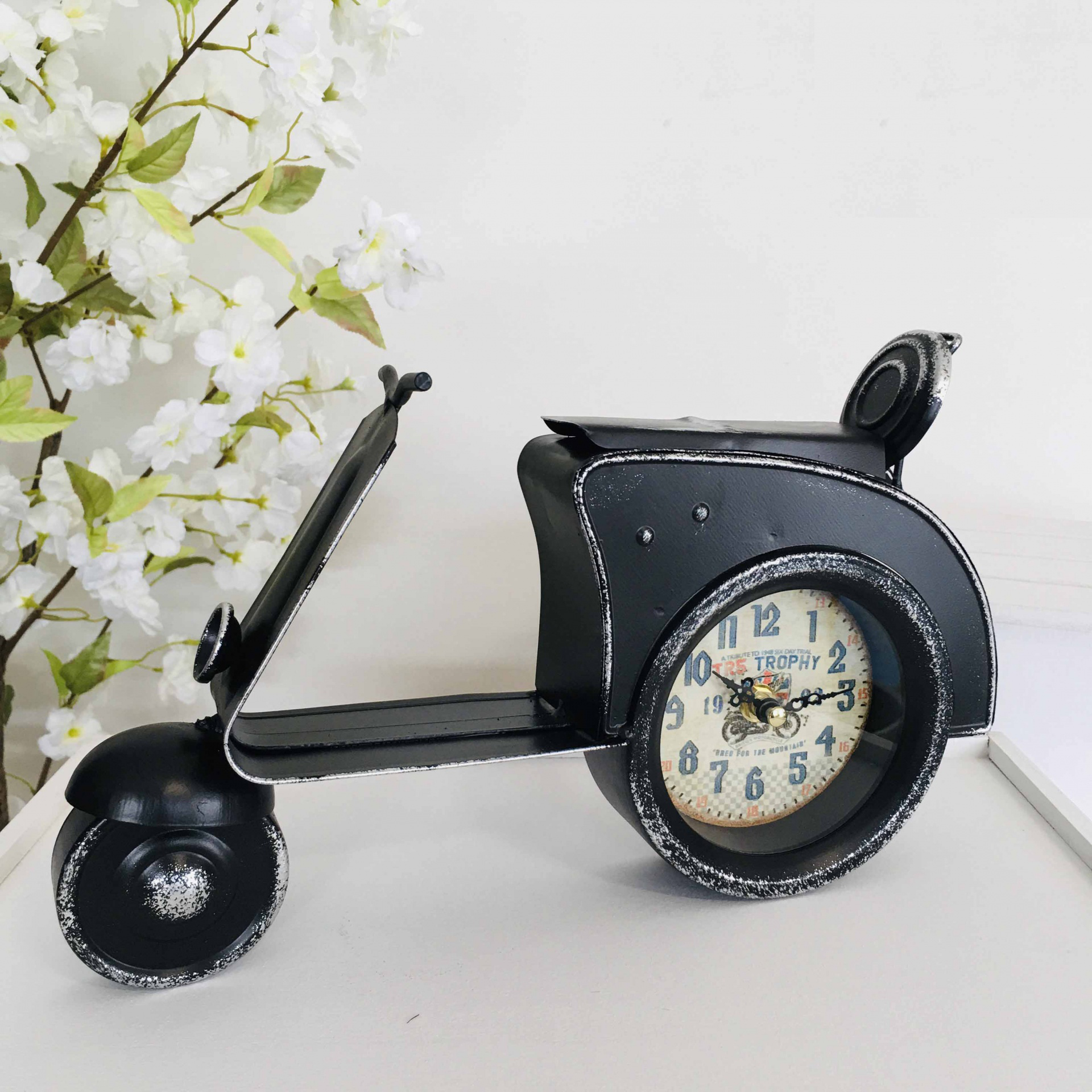 Relógio em formato de scooter vintage preta com mostrador redondo na roda sobre mesa branca próximo a flores brancas