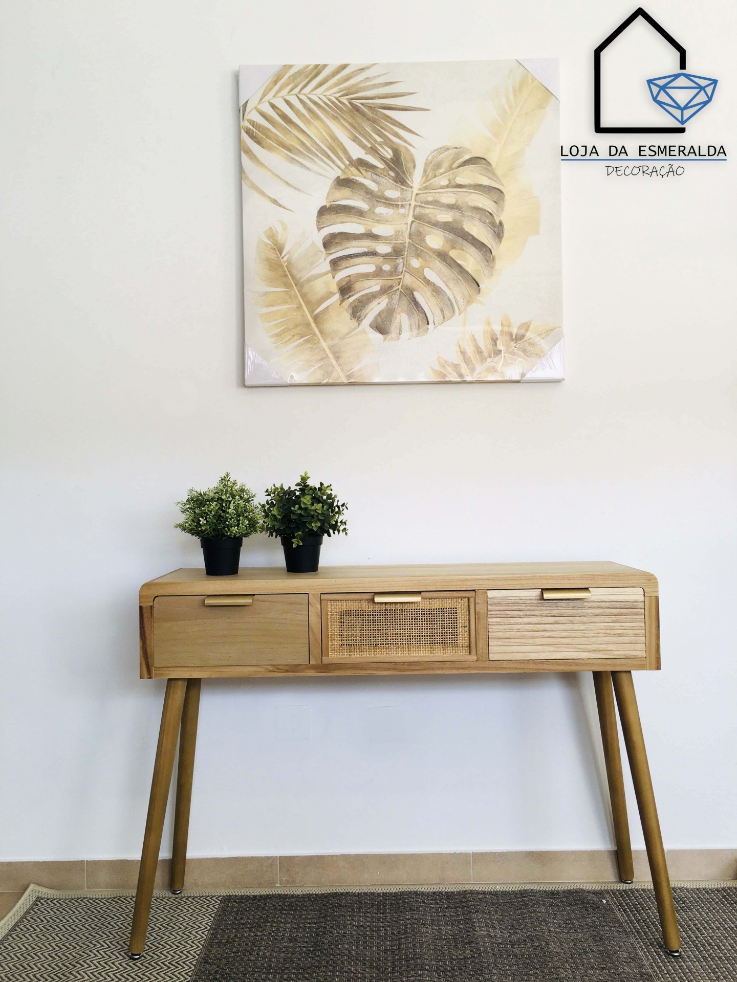 Mesa de madeira clara com três gavetas e plantas artificiais, com quadro de folhas douradas na parede