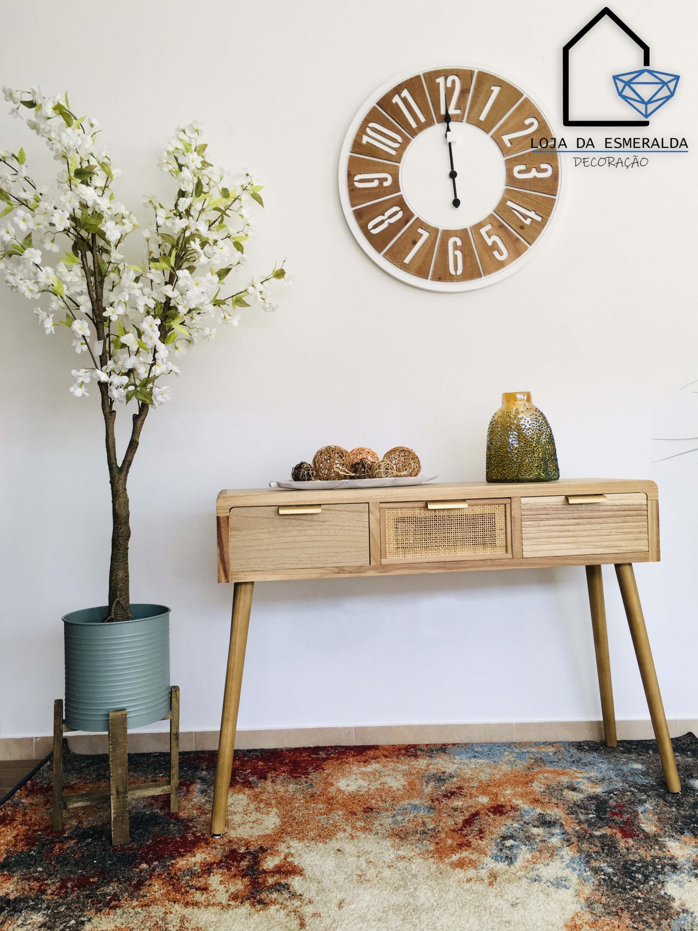 Relógio de parede de madeira, mesa de madeira clara com vaso e planta artificial, tapete colorido, texto LOJA DA ESMERALDA DECORAÇÃO