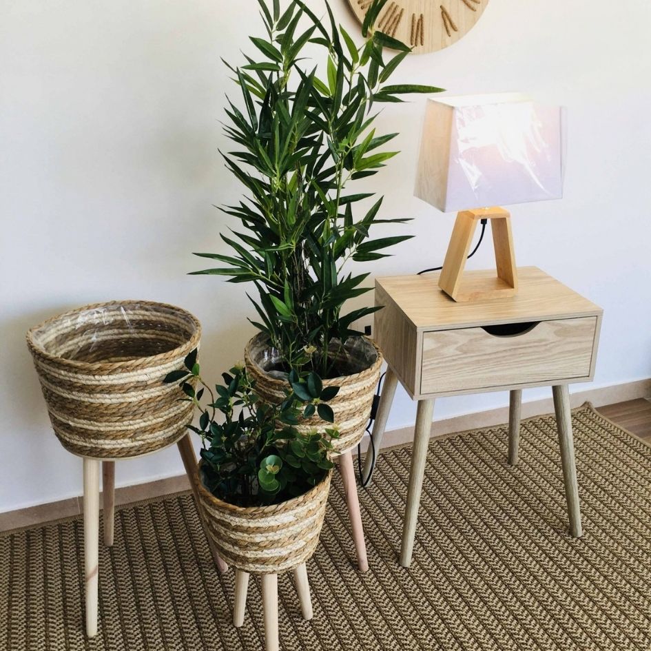 Floreiras de palha com plantas ao lado de mesa de cabeceira de madeira clara com lâmpada, sobre tapete castanho trançado