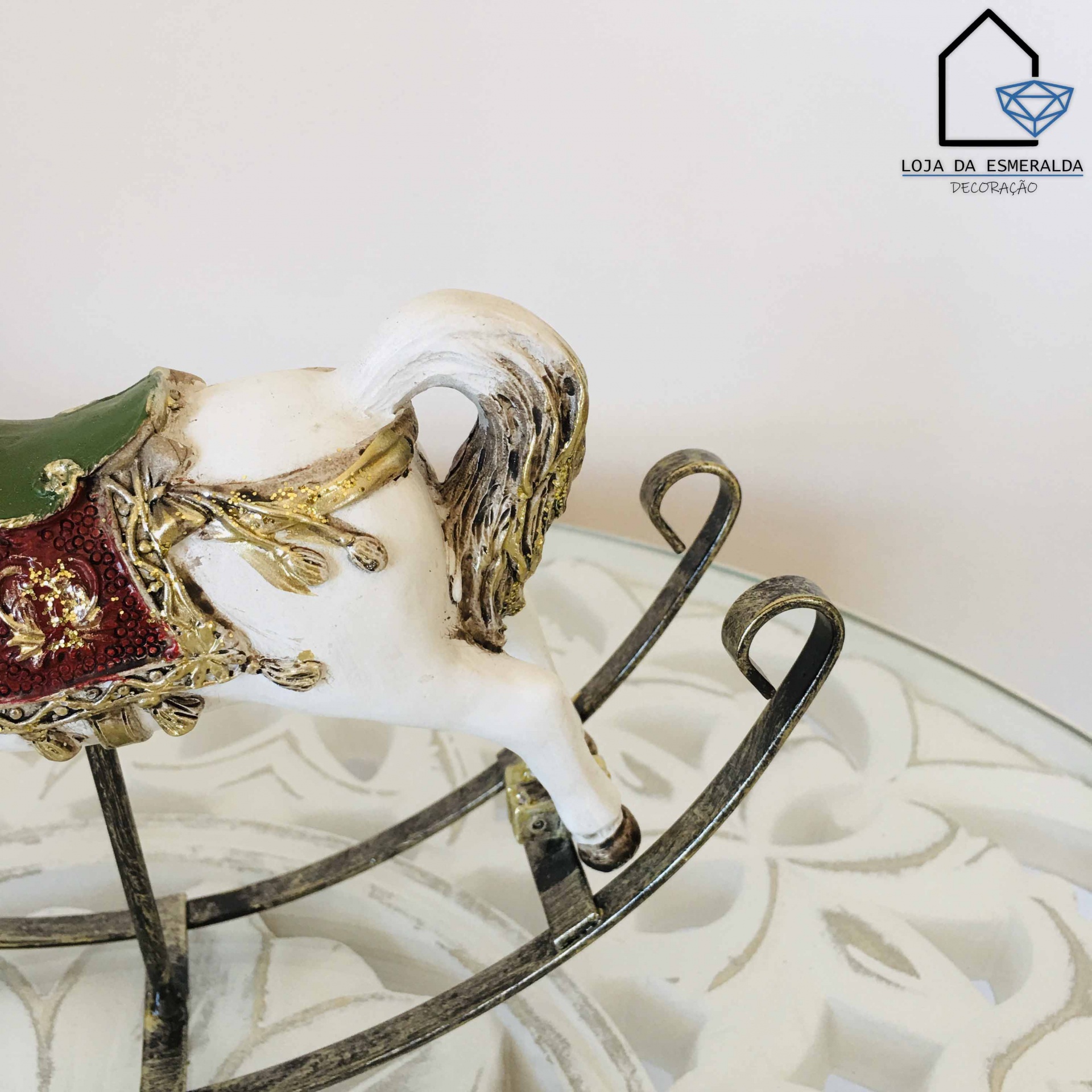 Cavalo baloiço decorativo branco com sela vermelha bordada e base metálica sobre superfície branca com padrão.
