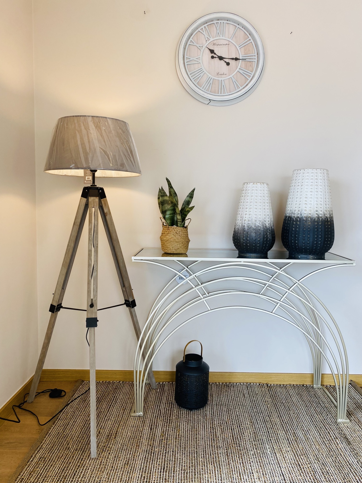 Relógio de parede, candeeiro de pé, mesa com vasos, lanterna e planta em ambiente interior