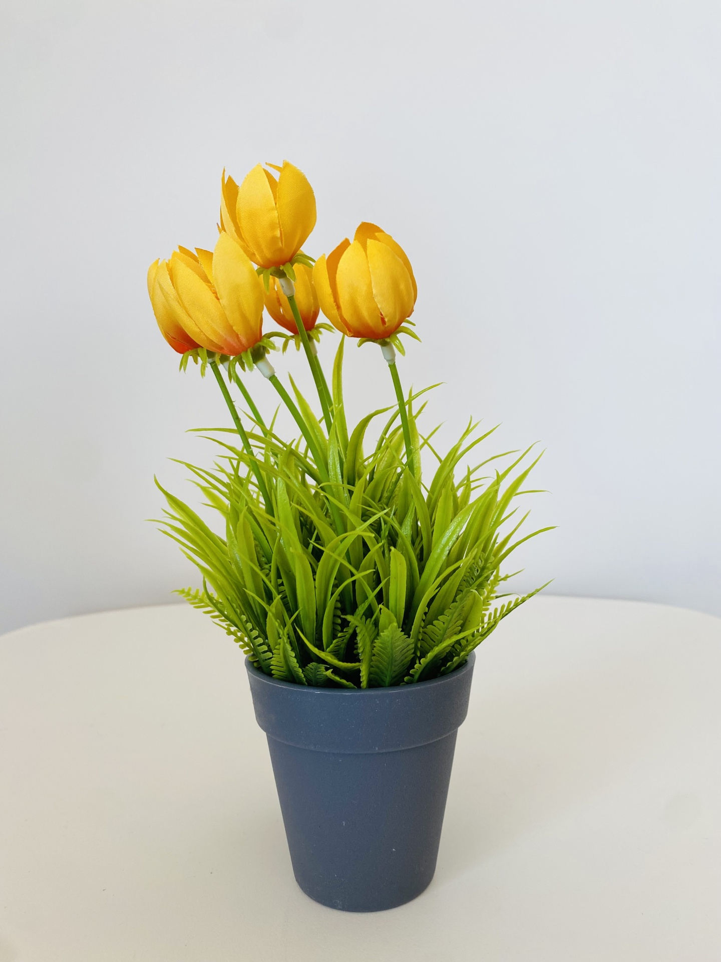 Vaso cinza com planta artificial de flores amarelas e folhas verdes em fundo branco