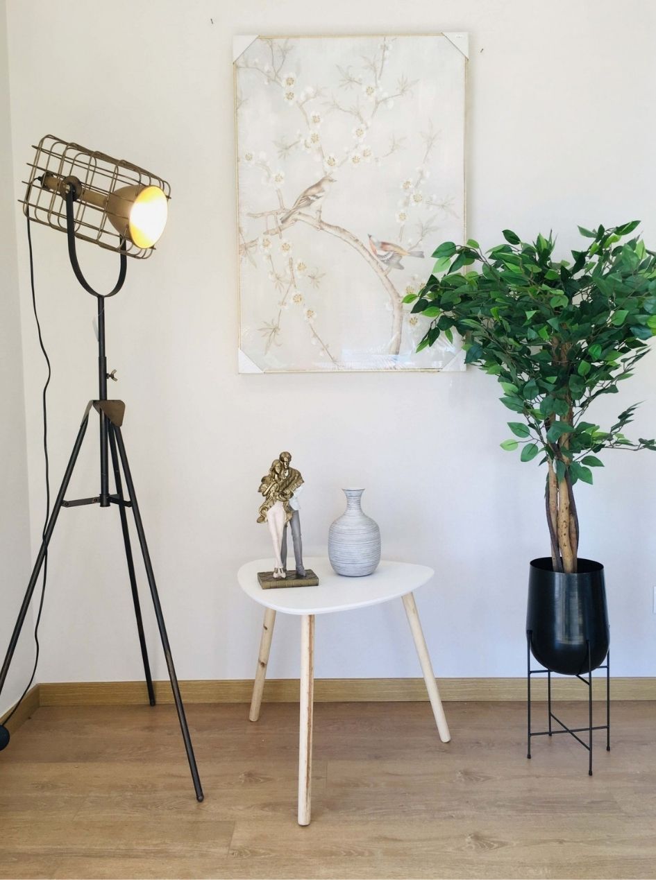 Conjunto de decoração com lâmpada de pé, mesa pequena, vaso, estatueta, quadro com ramo e pássaros e planta em vaso preto no chão.